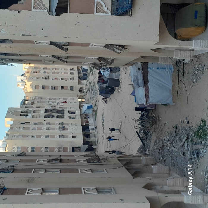 The image displays a severely damaged urban residential area in Khan Yunis, Palestinian Territory, under a clear sky. Multiple multi-story, light-colored apartment buildings, exhibiting extensive structural damage including numerous impact holes, broken windows, and compromised facades, frame an open, unpaved central ground. This central area is strewn with dirt and construction rubble, containing several makeshift shelters constructed from tarpaulins, fabrics, and metal debris; one shelter prominently displays a UNICEF logo. Laundry is visible hanging near some of these temporary structures. Approximately seven individuals, primarily appearing as adult males or youths, are present in the open space. Three individuals are clustered in the mid-ground, one with arms outstretched, while others are walking or standing. A vehicle, covered by a green and yellow tarp, is parked beneath an archway on the ground level of a damaged building in the lower right. Electrical wires are visible suspended across the scene.