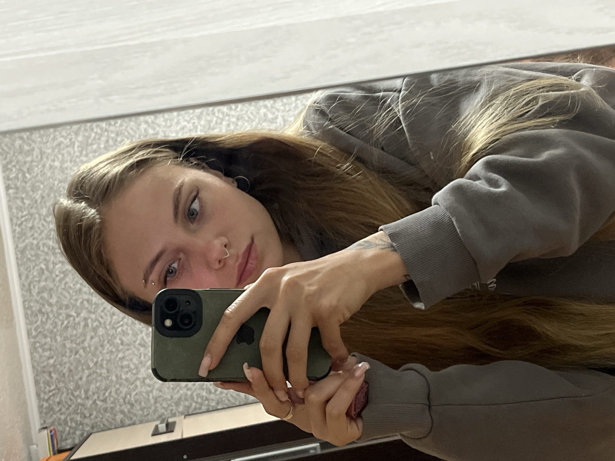 A young woman with long, blonde hair takes a selfie in a mirror. She is wearing a grey hoodie and has a nose ring and an eyebrow piercing. She is holding a dark green phone in her left hand. The background appears to be a bedroom with a closet and patterned wallpaper. The lighting is dim. The image was taken in Fokino, Russia.