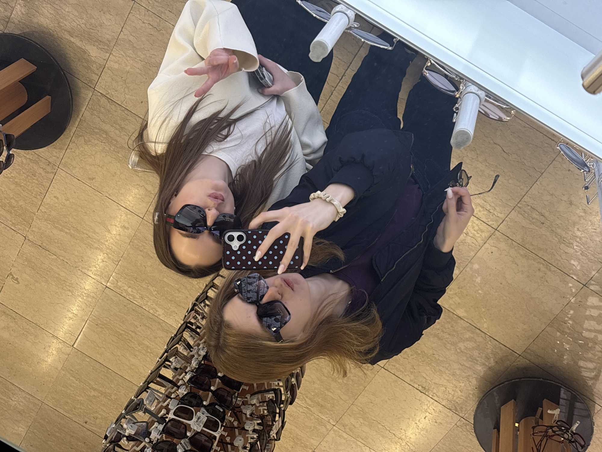 The image depicts two young women, reflected in a large mirror within what appears to be an optical store or sunglasses shop. They are engaged in taking a selfie.
The woman on the left, with long dark brown hair, wears a light-colored (off-white or cream) jacket over a white top and dark pants. She has dark, square-framed sunglasses with a distinctive red and green stripe detail on the temples. Her right hand is raised, making a peace sign, and her lips are slightly parted.
The woman on the right, with medium brown hair, is wearing a dark, puffy jacket over a purple top and dark pants. She holds a black smartphone with a dark case featuring light blue/white polka dots in her right hand, seemingly capturing the image. She wears patterned, dark sunglasses, and a light-colored scrunchie is visible on her left wrist. Her left hand also holds another pair of sunglasses by the arm.
The background reveals numerous sunglasses displayed on racks or stands, both in the foreground (bottom left) and background (top right, reflected on what appears to be a ceiling or upper wall fixture). Many display sunglasses have small white tags attached. The floor, visible through the reflection, is covered in light-colored square tiles. A black circular display stand with wooden inserts is visible on the far left and far right sides of the reflection.
The bright, even lighting suggests an indoor commercial environment. The overall mood is casual and possibly playful. Aside from the polka dots on the phone case, no other text is clearly legible within the image. The women's manicured fingernails are visible, particularly those of the woman holding the phone, which have a light polish and a subtle design. The scene gives the impression of friends shopping for and trying on sunglasses.
