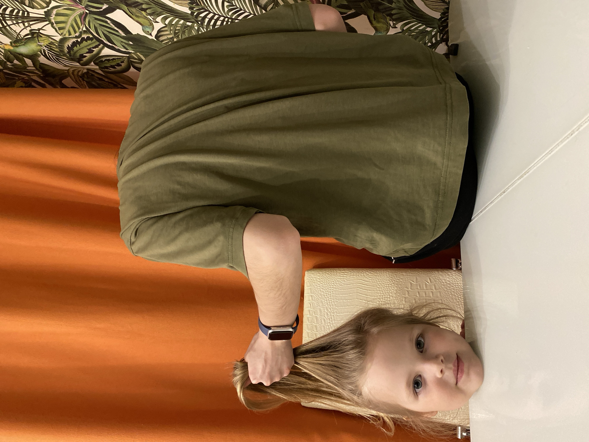 A horizontally oriented image, which appears to be rotated 90 degrees counter-clockwise, depicts an indoor scene involving an adult and a young child in Votkinsk, Russia.
On the left side of the frame, an adult is partially visible, wearing a dark olive green long-sleeved shirt. Their right arm is extended, with a smartwatch featuring a blue band and a silver-colored face on the wrist. The adult's hand is gently holding a lock of the child's light blonde hair.
On the right, a young girl with light blonde hair and blue-gray eyes is prominently featured. She looks directly at the camera with a calm and direct expression. Her head is resting on a smooth, white surface, likely a table or desk, which extends from the lower right of the frame. Behind her head, a light-colored, possibly beige, object with a textured, crocodile-skin-like pattern is visible, possibly a cushion or a chair back.
The background is composed of two distinct elements. The upper part features wallpaper adorned with a vibrant tropical pattern, showcasing large green leaves and hints of birds, possibly parrots or toucans, against a lighter background. The lower portion of the background is dominated by a pleated orange or burnt orange curtain, providing a warm color contrast to the scene. The lighting is soft and even, typical of an indoor setting with diffused light. No text is visible in the image.