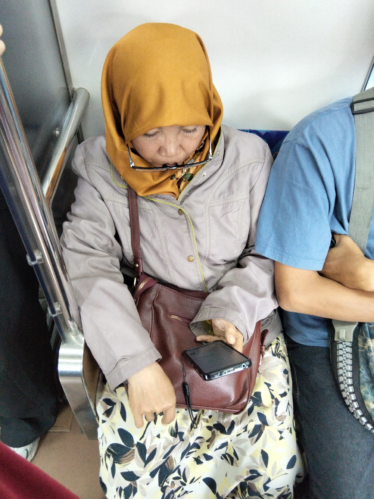 A woman wearing a mustard-yellow hijab and glasses sits on a blue seat, her head bowed towards a smartphone she holds in her right hand. She is wearing a light grey jacket over a floral-patterned dress and a brown purse hangs from her shoulder. To her right, another person in a blue shirt sits with their arms crossed. The scene appears to be on public transportation, possibly a bus, given the presence of a metal railing and upholstered seats. The lighting is consistent with an indoor environment. The image is taken from a slightly elevated perspective, looking down at the woman.