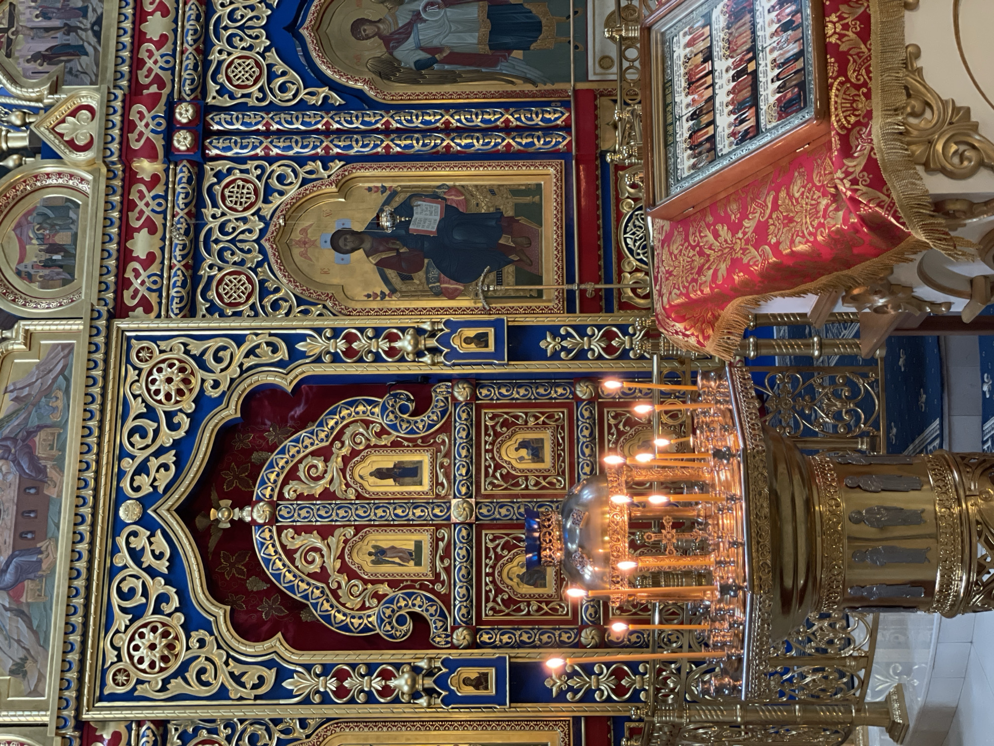 The image shows an interior view of a religious building, likely a church, in Krasnoyarsk, Russia. The architecture is ornate, featuring a red, blue, and gold color scheme. Religious icons and paintings adorn the walls, including depictions of religious figures. A large, multi-tiered candlestick is visible in the foreground, with many lit candles. The overall environment suggests a space of worship or religious significance.