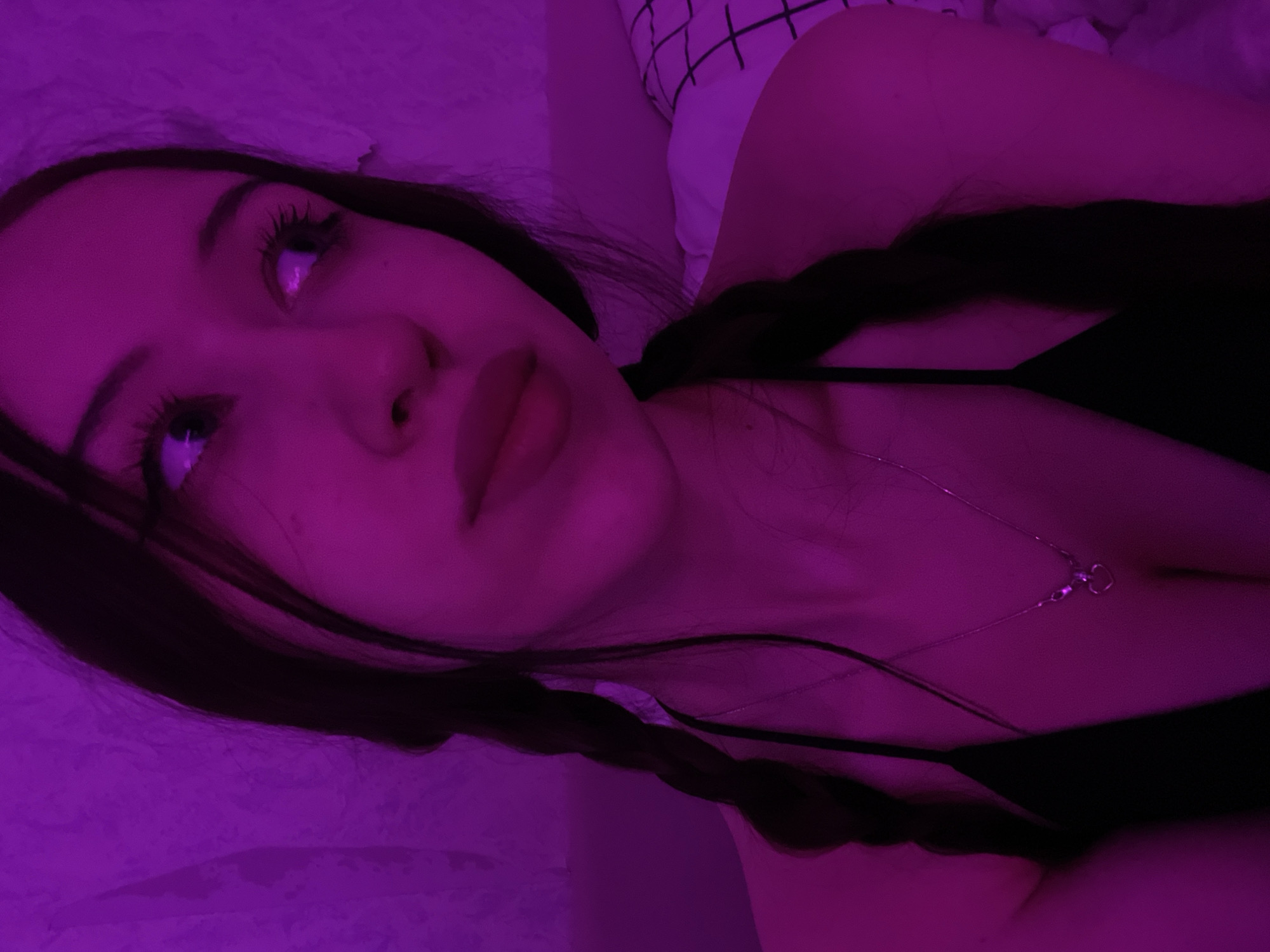 A young woman with dark hair in braids is looking away from the camera, illuminated by purple light. She is wearing a black top and a silver necklace with a heart pendant. The background is a textured wall. The image appears to be taken indoors. The location in Votkinsk, Russia, is not discernible from the image itself.