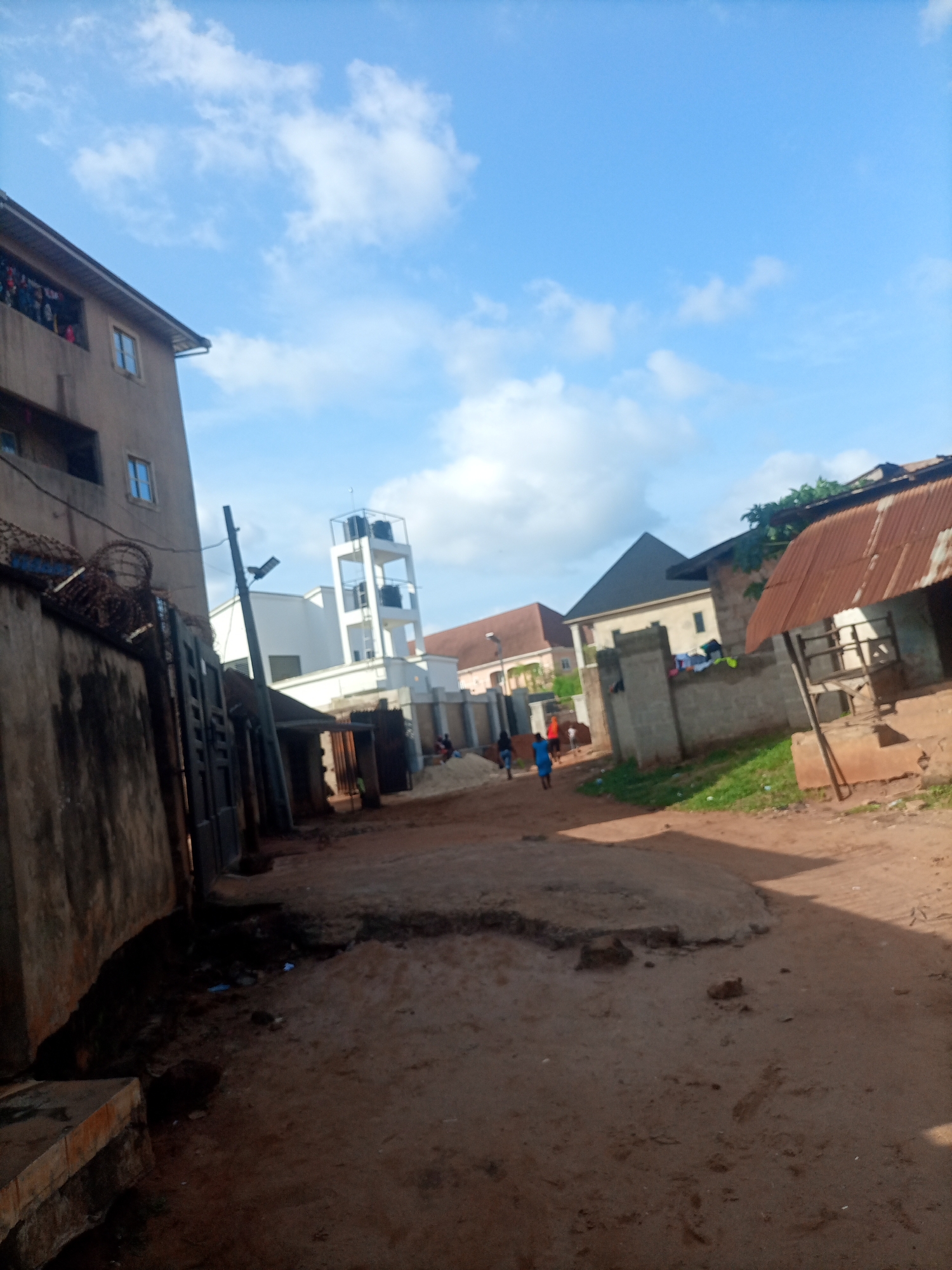 The image depicts a street scene in Nkpor, Nigeria, under a bright, partly cloudy sky, suggesting it is daytime. The street is unpaved, with areas of packed dirt and some uneven, raised patches. To the left, a multi-story building with balconies and barbed wire fencing is visible. A large concrete wall runs along the foreground.
In the middle ground, a modern-looking white building with a water tower structure is prominent. Further down the street, several other buildings are visible, including one with a corrugated metal roof and another with a dark, pyramidal roof. There are a few people in the scene, some walking or standing, and others appear to be in the background near the buildings. Laundry is hanging on a wall, suggesting residential activity. The overall impression is of a typical urban or semi-urban environment in Nigeria, with a mix of older and newer structures and evidence of daily life. No visible text is present in the image.