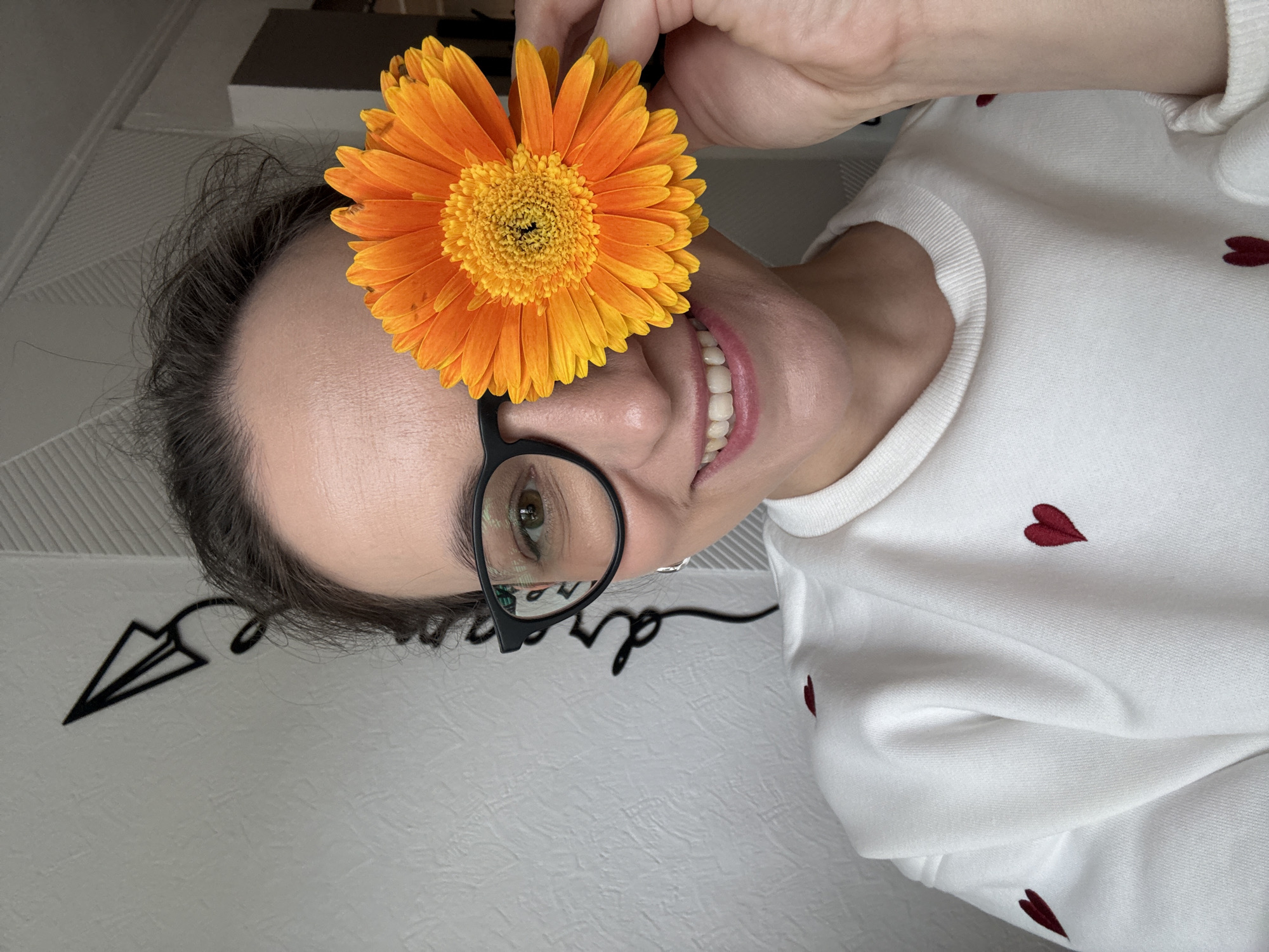 A woman is holding an orange gerbera daisy flower in front of her face. She is wearing black-rimmed glasses and a white sweater with small red heart embroidery. The background is a light-colored wall with a decorative paper airplane design. The image does not contain sufficient information to determine the location in Chaikovsky, Russia.