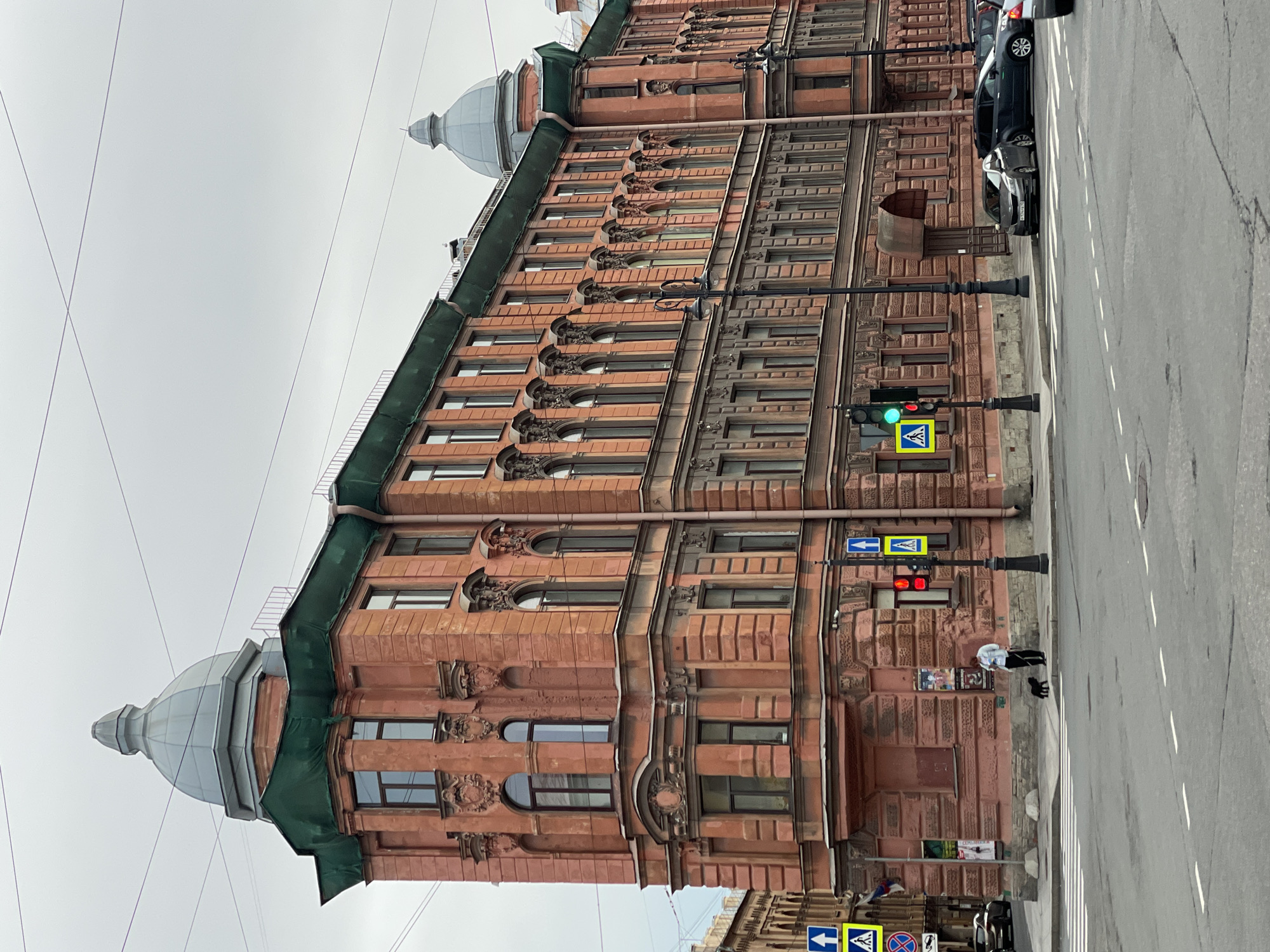 The media file shows a tilted, wide-angle view of a historic building with terracotta-colored facades on a street in Saint Petersburg, Russia. The building features ornate architectural details, including arched windows, decorative carvings, and two prominent domed turrets. Electrical wires crisscross the sky, and a traffic light with a green signal is visible. Cars are parked along the street, and a person is walking with two dogs on the sidewalk. The weather appears overcast, suggesting a cloudy day. The overall impression is one of a bustling urban environment with a rich architectural heritage.