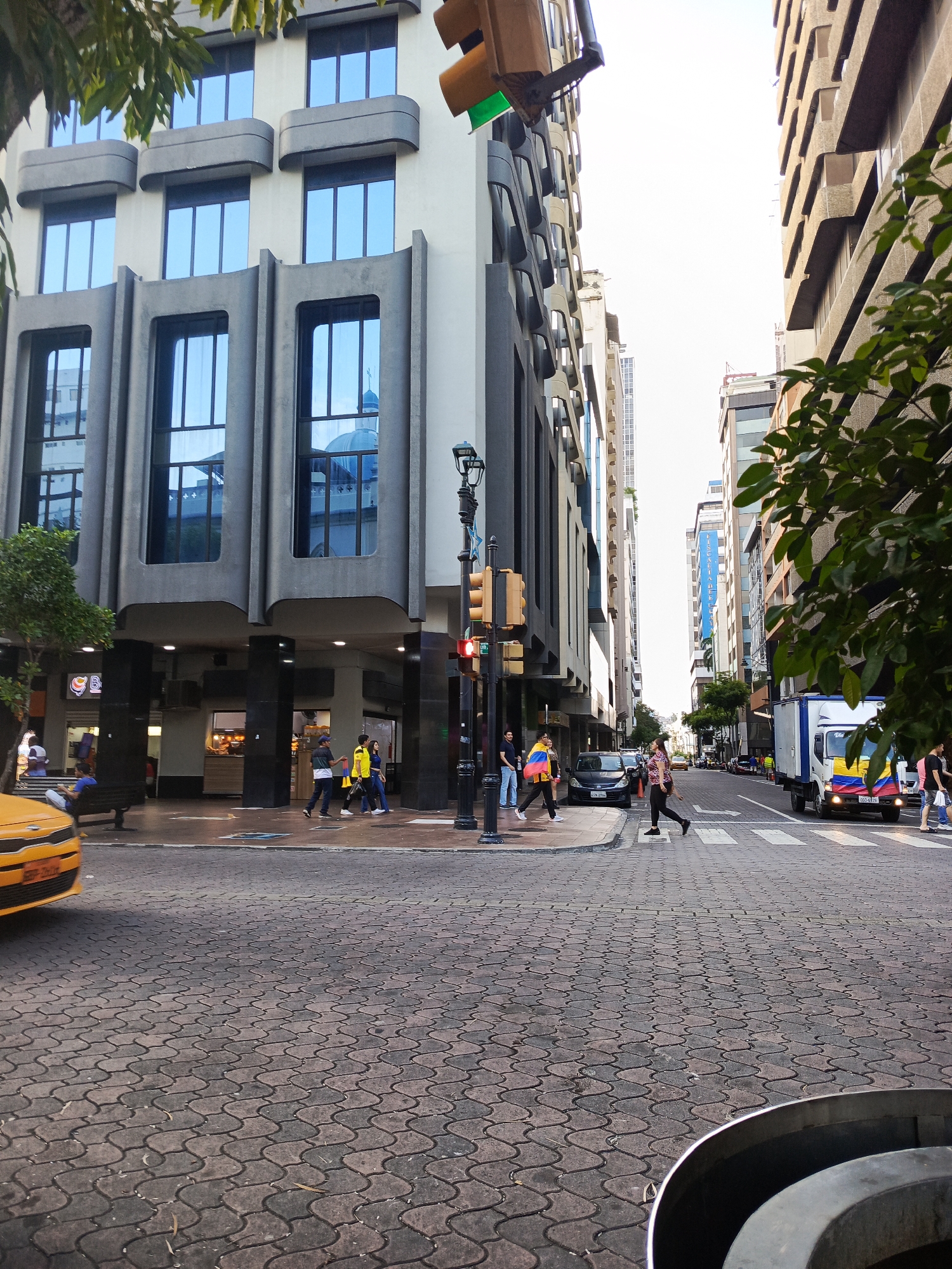 The image captures an urban street scene in Guayaquil, Ecuador, likely during daylight. The perspective is from street level, looking down an intersection paved with decorative interlocking bricks. Tall, modern buildings with reflective glass windows dominate the cityscape, their facades varying in architectural style.
On the left, a building features a striking concrete facade with curved elements and large, recessed windows. A yellow taxi is partially visible in the foreground. People are walking on the sidewalk and crossing the street at a marked crosswalk.
On the right, another building rises, its facade characterized by repeated horizontal concrete structures. A white truck with a colorful flag design, possibly representing Ecuador, is seen at the crosswalk. Pedestrians are also visible on this side of the street.
Traffic lights are present, with one showing a red light. Trees with green foliage can be seen, suggesting the presence of street greenery. The sky is bright, indicating it is likely daytime. Reflections of buildings and a dome-like structure, possibly a church or government building, are visible in the windows of the building on the left. The overall impression is a busy, contemporary urban environment.