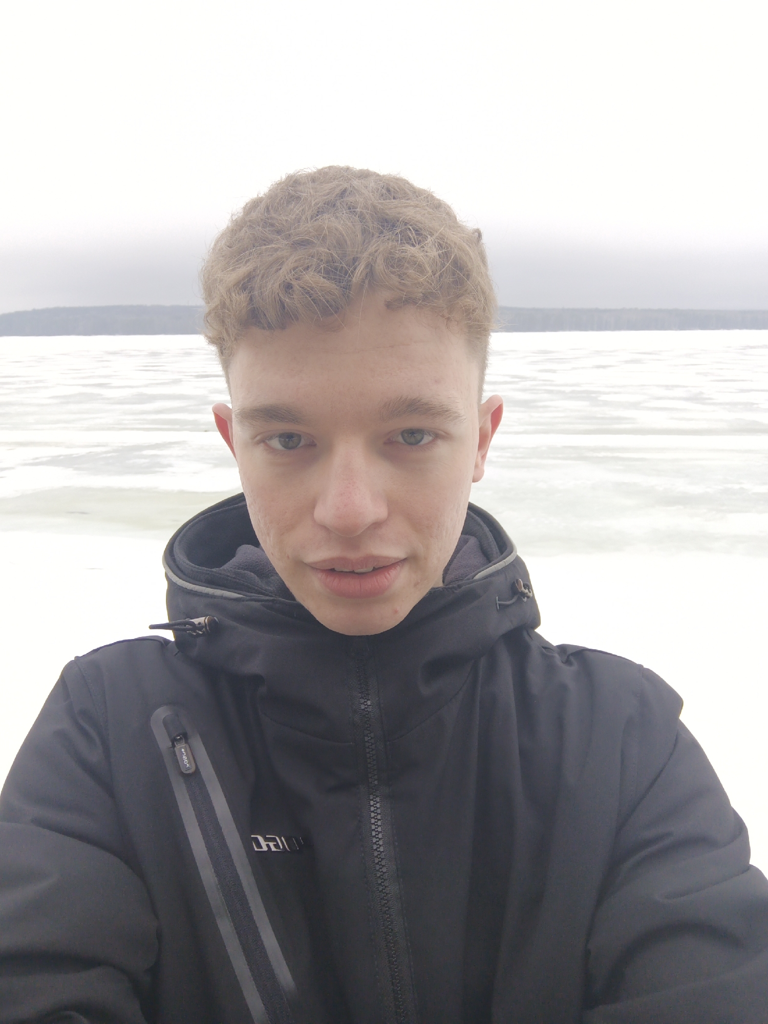 A young Caucasian man is facing the camera, wearing a black winter jacket. Behind him, there is a frozen body of water, possibly a lake or a river, with ice and snow covering its surface. A line of trees is visible in the background beneath a dull, overcast sky. The location is likely in Chaikovsky, Russia.