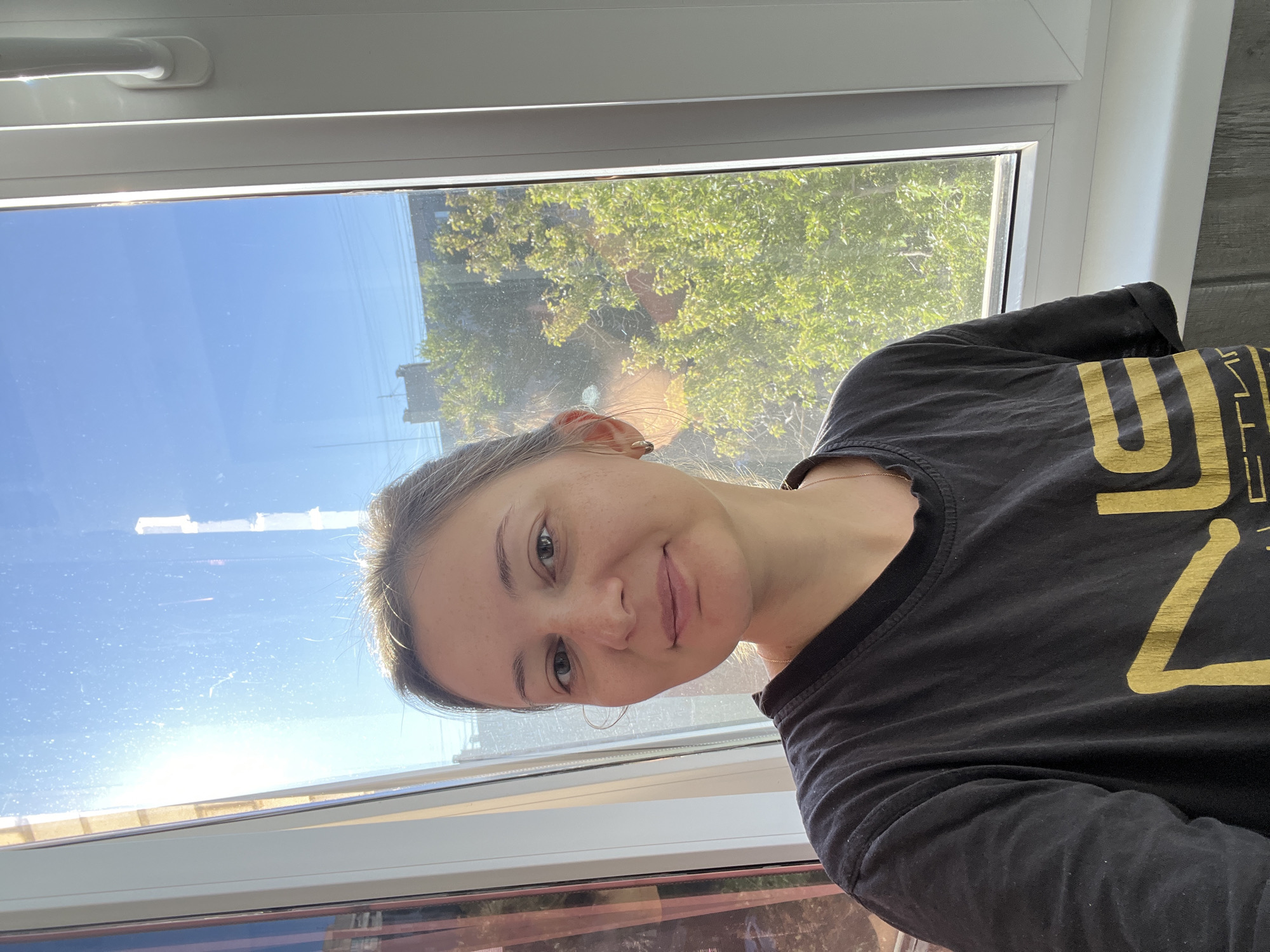A medium shot captures a young adult woman with light skin, dark brown hair pulled back, and light green eyes, looking directly at the camera. She is wearing a black t-shirt with a gold graphic design. The woman is positioned in front of a window with a white frame, which overlooks a sunlit urban landscape. Lush green trees are visible through the window, alongside buildings and a bright blue sky. The light streaming through the window creates a lens flare effect. The scene appears to be indoors, likely in an apartment, given the window and interior wall details visible on the right. The overall impression is a casual, daytime photograph taken indoors. Volgograd, Russia.