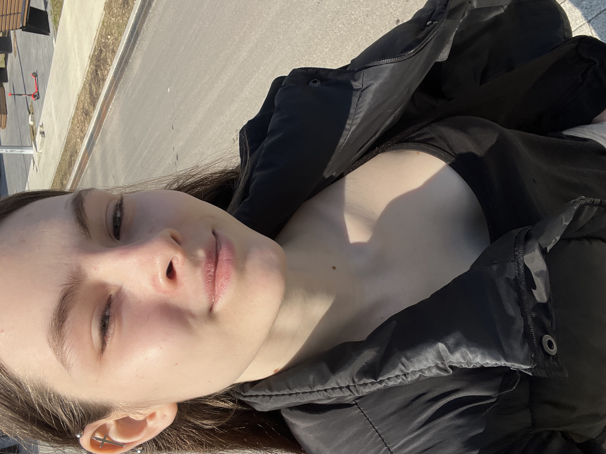 A young woman lies on her back outdoors. She is wearing a black down jacket over a black top. The location appears to be an urban area, potentially in Saint Petersburg, Russia, based on the architectural style and landscaping in the background, visible in the upper-left corner (gray pavement, a stretch of green space with thin grass and bare trees, and a red scooter). The angle of the shot is from above, close to the woman's face, which is partially in sunlight. Her eyes are mostly closed, suggesting she is relaxing or sunbathing. She has an industrial piercing with a cross on her left ear.