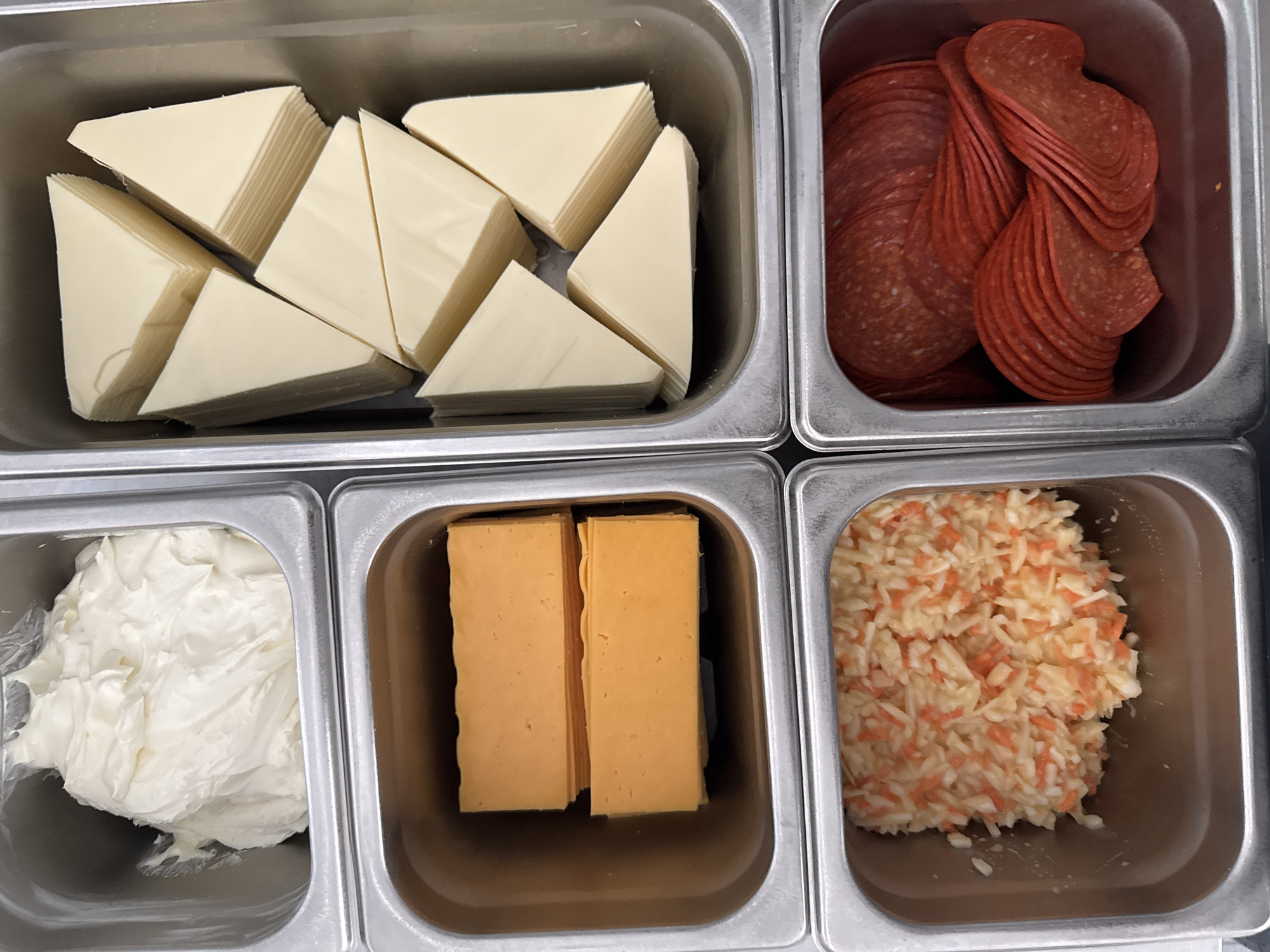 An overhead shot shows four stainless steel food pans arranged in a two-by-two grid. The top left pan contains a mound of creamy white substance, possibly cream cheese. The top right pan is filled with stacked, triangular slices of pale yellow cheese. The bottom left pan holds two rectangular blocks of bright orange cheese. The bottom right pan contains finely shredded coleslaw, a mixture of white and orange shreds. In the bottom right, there is a pan of sliced pepperoni. The image was taken in an unknown location.