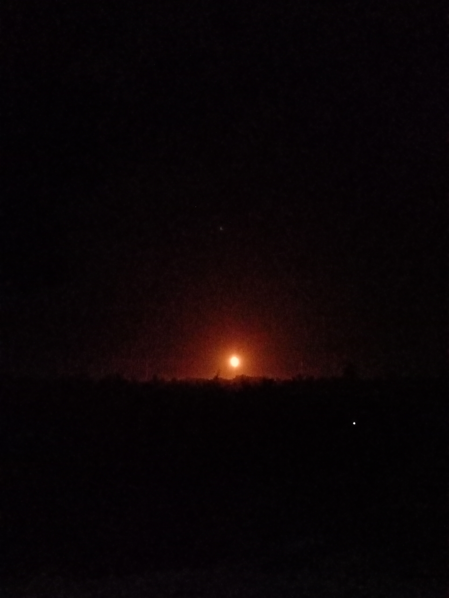 The image shows a very dark scene with a bright, orange-red light source near the horizon, likely the setting sun. A dark, undefined land mass, possibly trees or a low hill, stretches across the horizon. There is a second, smaller light point visible in the upper half of the image. Due to extreme darkness and lack of detail, it is difficult to determine a more precise location within Lhokseumawe, Indonesia, or to describe any subjects or activities.