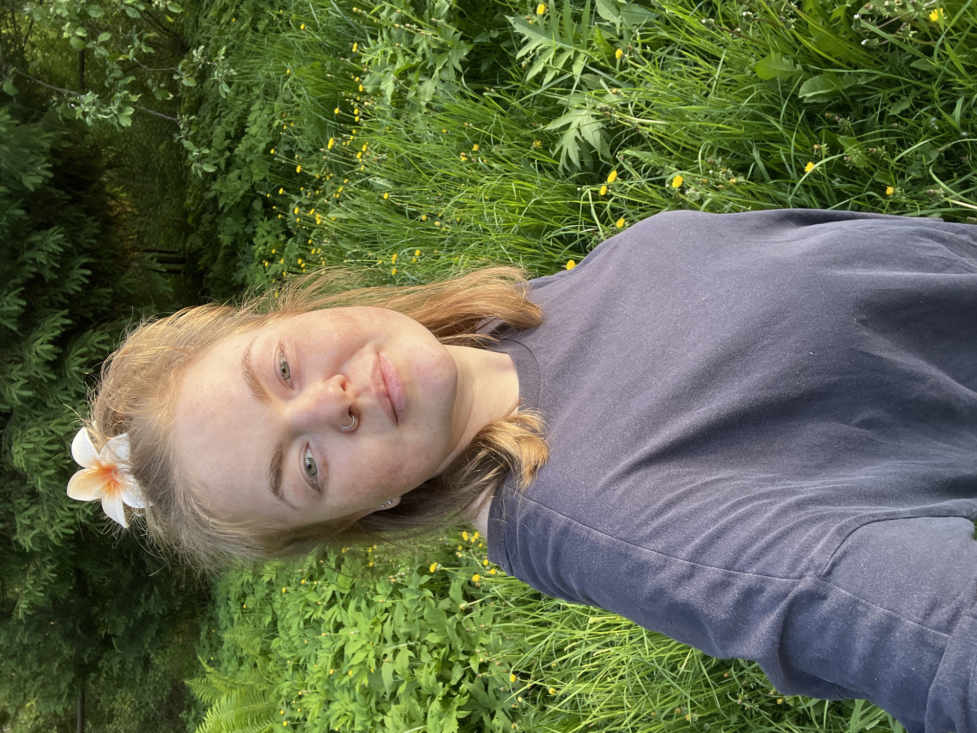 A young woman lies in a grassy area with scattered yellow wildflowers. She has light blonde hair, fair skin, and a nose ring. A white and orange flower is tucked into her hair. She wears a dark blue or grey T-shirt. The background consists of more grass and a section of trees. There is no specific information in the image to confirm or deny that this is in the city of Chaikovsky, Russia.