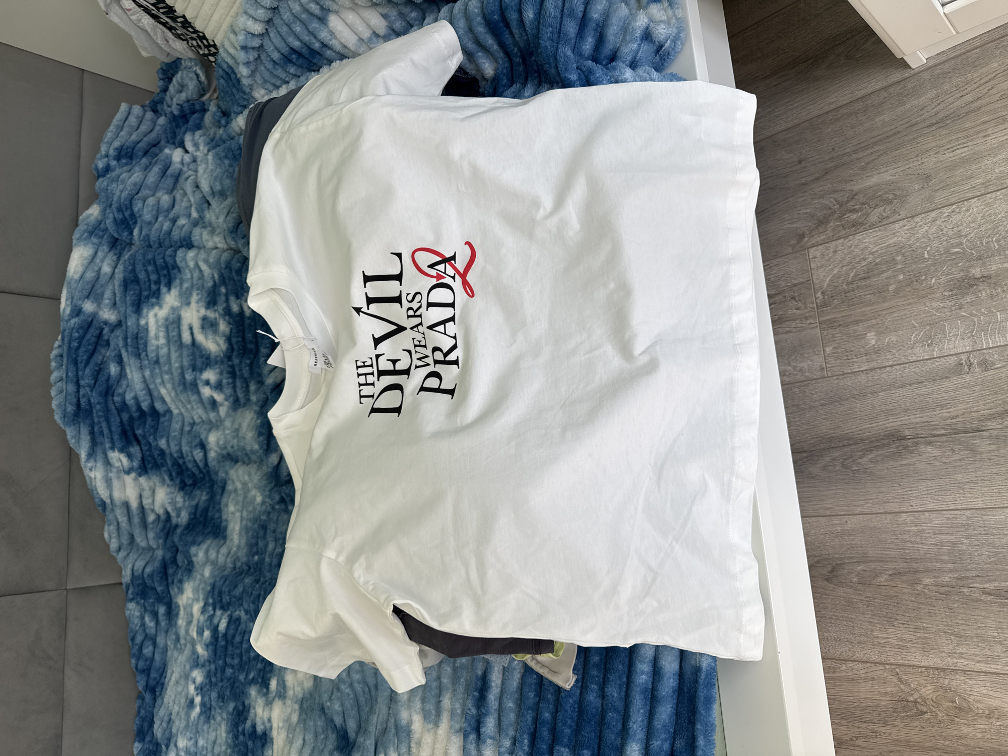 A white t-shirt with the text "THE DEVIL WEARS PRADA" in black and red lettering is lying on top of a blue and white patterned blanket. The blanket is on a gray surface, possibly a sofa or bed. The background includes a light wood floor and a white drawer. The scene is indoors. It is not possible to determine if the location is in Sofia, Bulgaria from the image.
