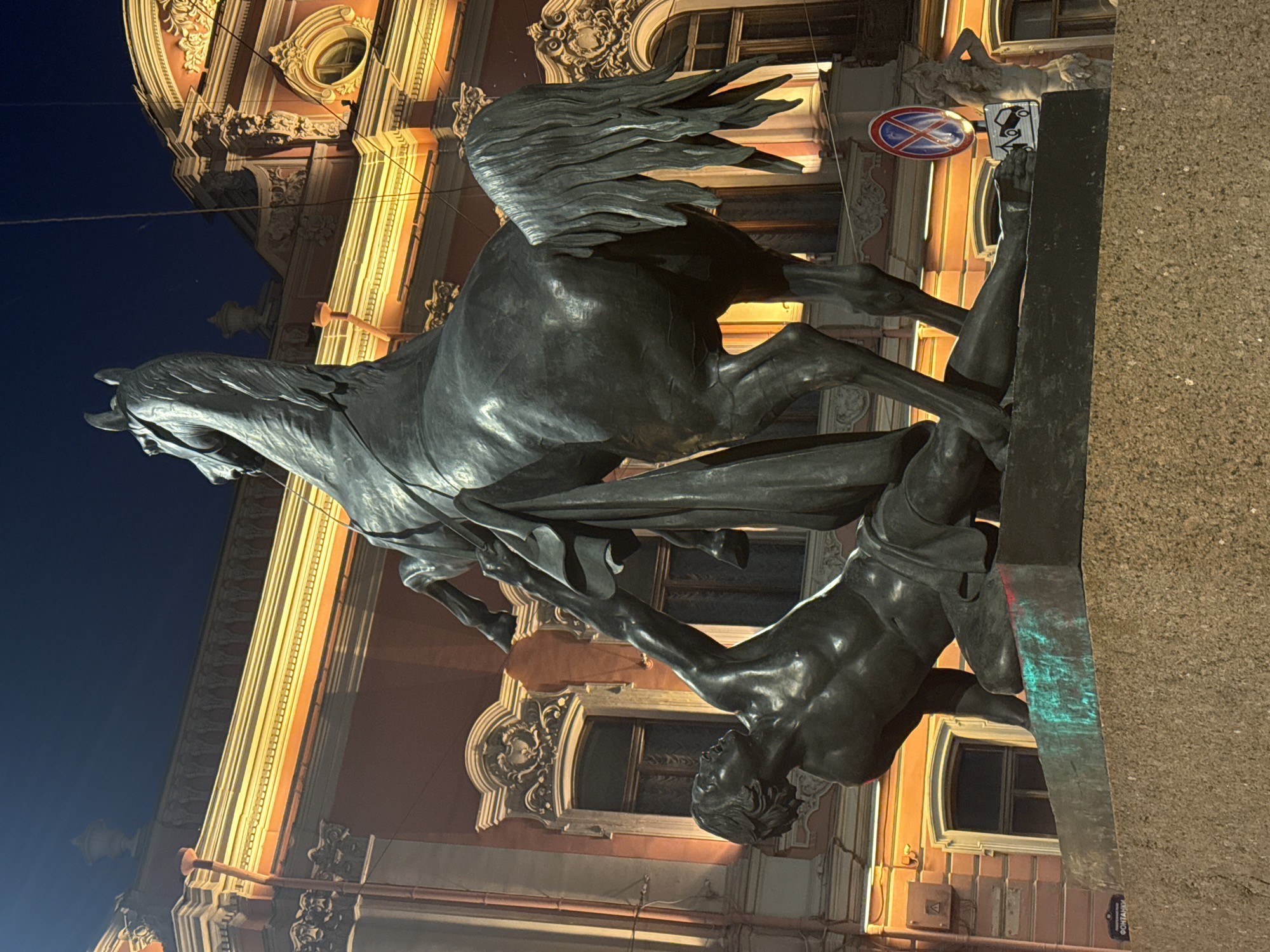 The image shows the Bronze Horseman, a monument to Peter the Great, located on Senate Square in Saint Petersburg, Russia. The statue, made of bronze, depicts Peter the Great on horseback, with the horse rearing up and trampling a serpent. The monument is illuminated by warm, orange lights from a building in the background, which is likely the General Staff Building. The sky above is dark, indicating it is nighttime. The statue is situated on a large granite pedestal, known as the "Thunder Stone," which was transported from Finland. In the foreground, part of the pedestal and some street signage are visible. The overall scene captures the grandeur and historical significance of the monument in its urban setting.