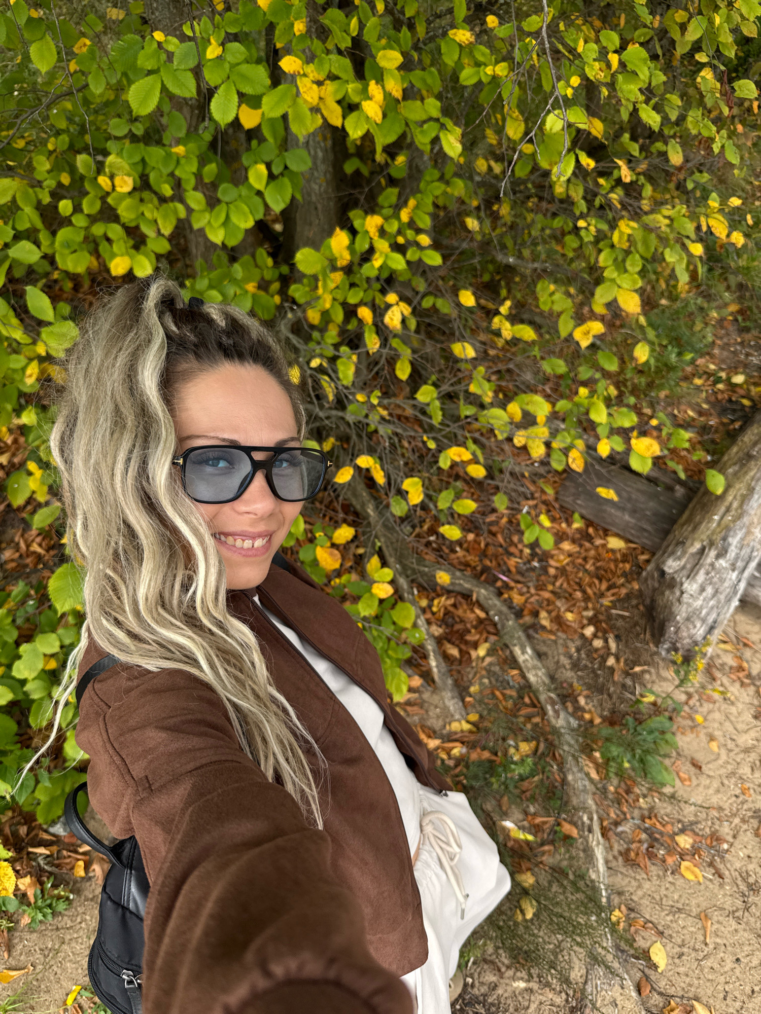 A woman with blonde hair pulled into a high ponytail, wearing large sunglasses and a brown jacket over a white top, is taking a selfie outdoors. She is smiling at the camera. The background consists of trees with green and yellow leaves, and fallen leaves on the ground, suggesting an autumn setting. The ground appears to be sandy or dirt. Visible objects include a black backpack on the woman's left side. The location is specified as Ignatovo, Russia.