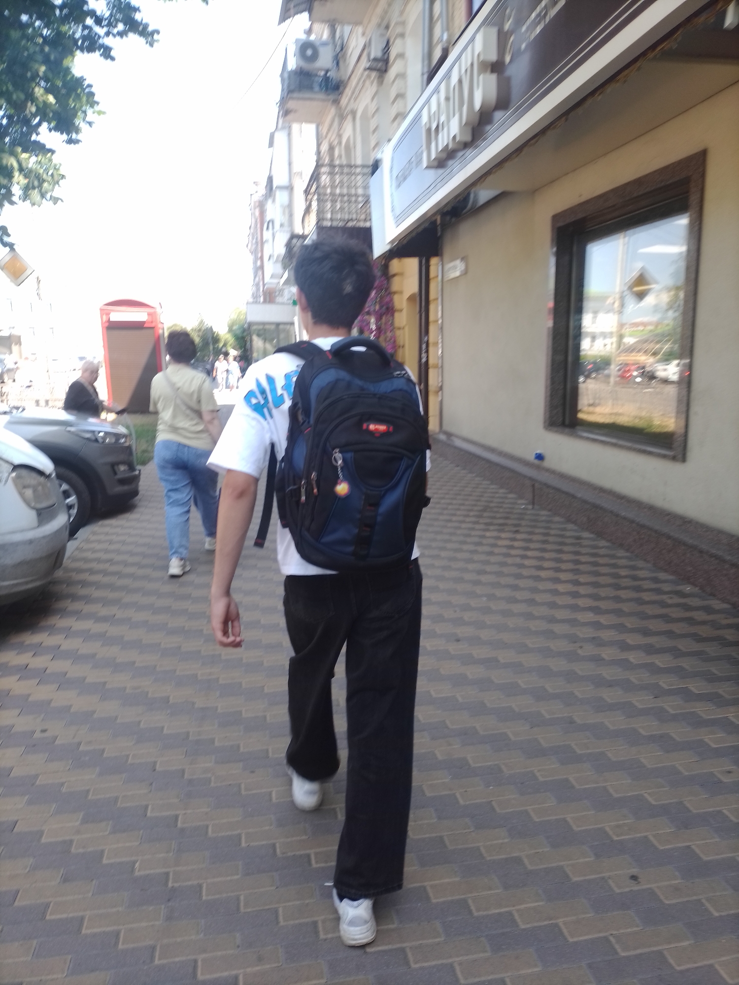 The image depicts a street scene in Poltava, Ukraine, with a focus on a young male with dark hair wearing a white t-shirt with blue writing and a dark backpack walking away from the camera down a paved sidewalk. He is wearing black pants and white sneakers. To his left, a woman with dark hair wearing a light-colored t-shirt and blue jeans is walking in the same direction, also at a distance. Further to the left, a man in a dark jacket is partially visible near a silver car. The sidewalk is made of interlocking brick pavers. In the background, there is a building with a shop facade featuring signage. One sign, partially visible, appears to be in Cyrillic. The sky is bright and blown out, suggesting it is daytime. A red structure, possibly a kiosk or booth, is visible in the mid-ground to the left.