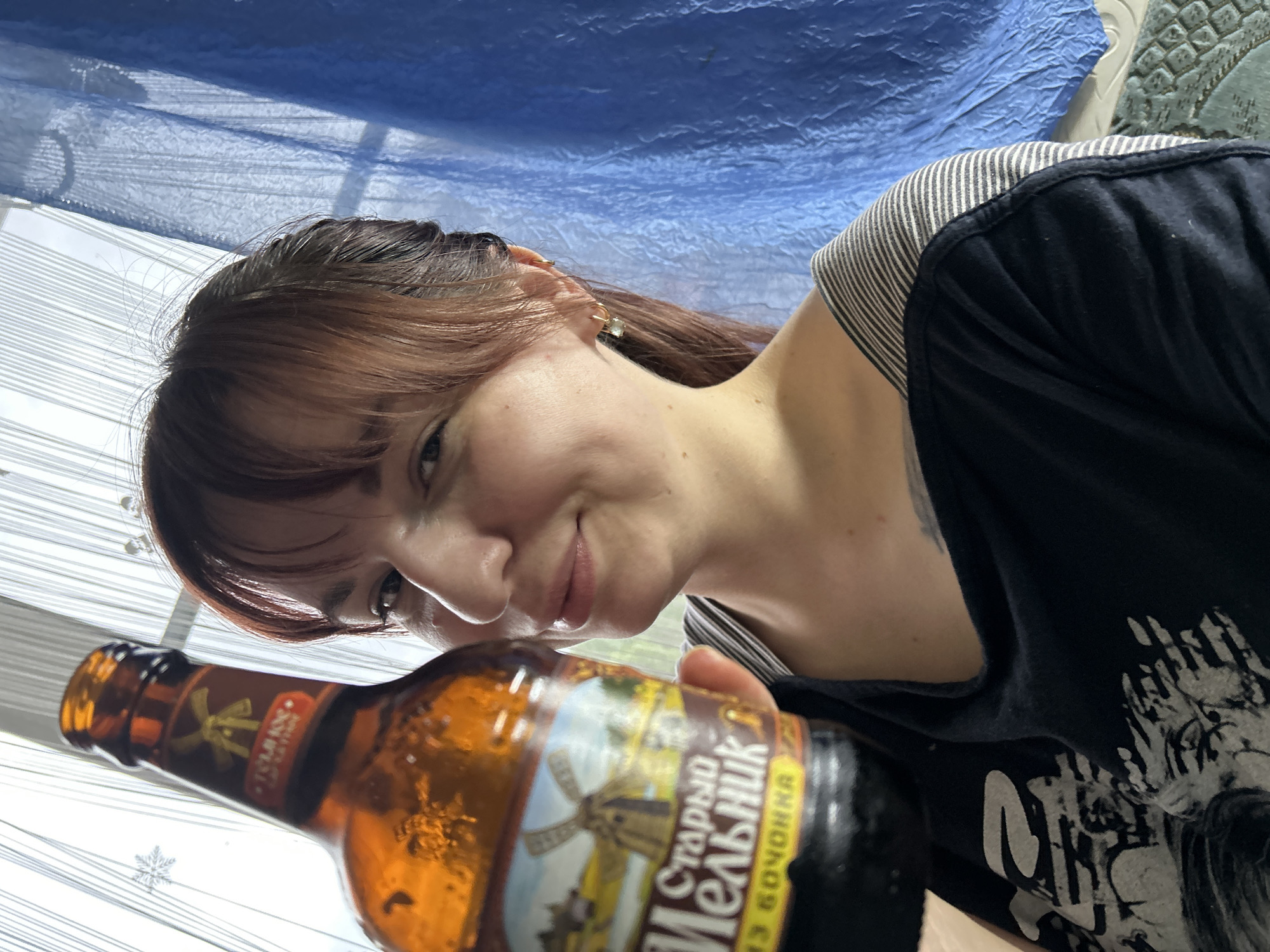 A female subject with light skin and dark brown hair, styled with bangs and pulled back, is depicted indoors in Фоки, Russia. She is wearing multiple small, gold-toned hoop earrings, one of which includes a clear square stone. Her attire consists of a black short-sleeved t-shirt with a light-colored graphic print on the front, layered over a horizontally striped (white and grey/blue) garment visible at the neckline. A dark tattoo is partially visible on her upper right chest/shoulder area. The subject is holding a brown glass beer bottle with her right hand, positioned in front of her. The bottle's main label features a windmill illustration and Cyrillic text reading "Старый Мельник" (Stary Melnik) and "ИЗ БОЧОНКА" (From the Barrel). The bottle's neck label also displays a windmill and "ТОМЛЁНОЕ" (Tomlyonoe). The subject is looking towards the camera with a slight smile. The background shows a window covered with vertical white blinds or sheer curtains, with a snowflake decal affixed to the left side. To the right, a blue draped fabric or tarp is present, covering part of the wall, with a textured floor or mat visible below it.