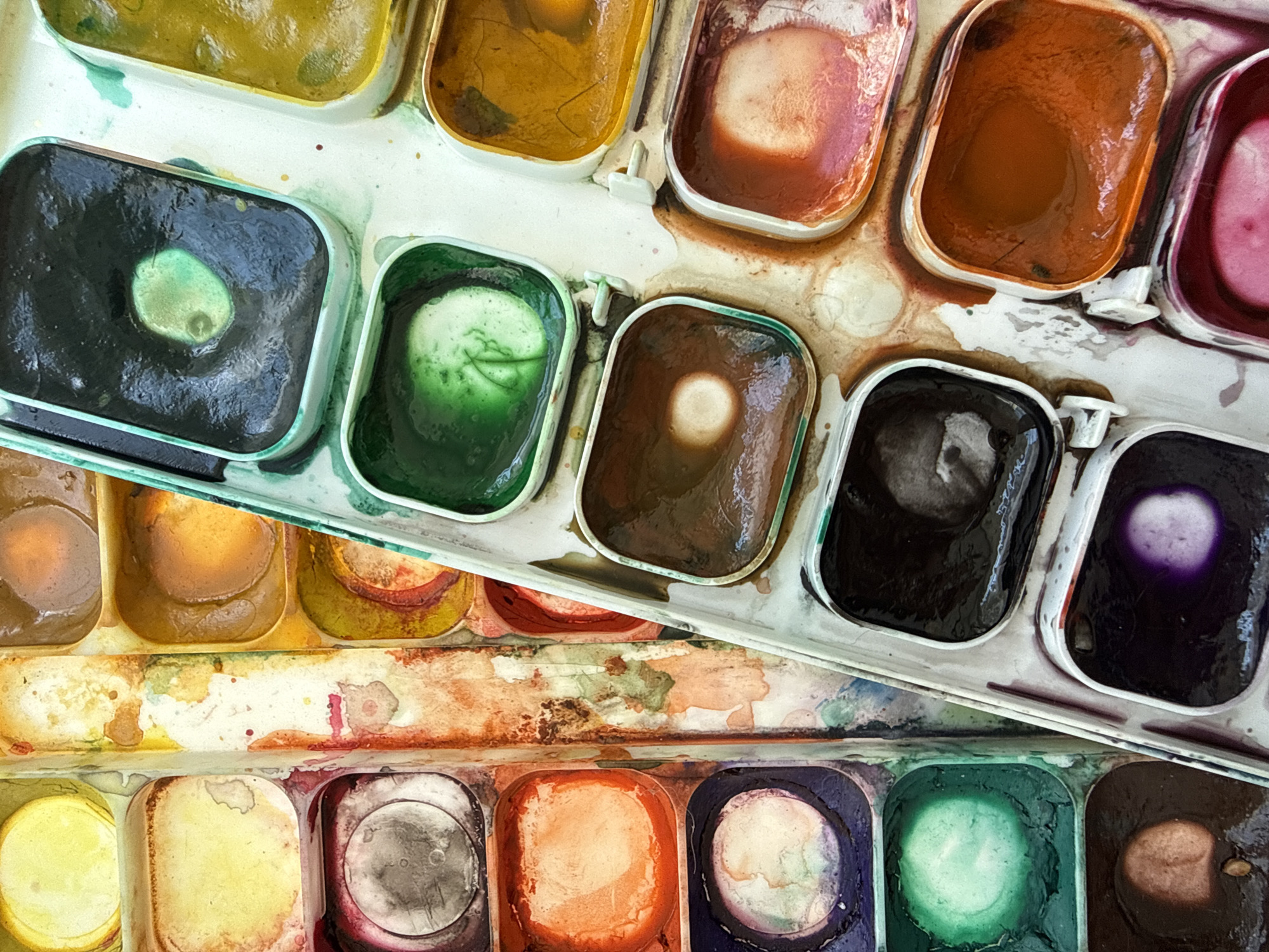 The image shows a close-up, overhead view of a well-used watercolor paint set. The paints are arranged in rectangular pans within a white plastic tray. The tray is tilted, revealing two rows of paint pans. The upper row has pans filled with various colors, including a deep black with a purple highlight, a dark brown, a rich green, a dark greyish-blue, and an orangey-red. Some pans show remnants of dried paint and water, with areas of lighter color where water has evaporated, leaving behind more concentrated pigment. The lower row, visible beneath the upper tray, contains more pans of paint, including yellow, orange, and a darker reddish-brown. The surface of the paint pans is uneven, with some areas appearing wet and glossy, while others are dry and matte. The white plastic tray is stained and splattered with paint, indicating its frequent use. There are no people, animals, or explicit location cues visible in the image, other than the provided metadata that this image is from Vladivostok, Russia. The lighting appears to be natural and diffused, suggesting an indoor setting, possibly an artist's studio or workspace. The focus is entirely on the art supplies, highlighting the texture and varied states of the watercolors.