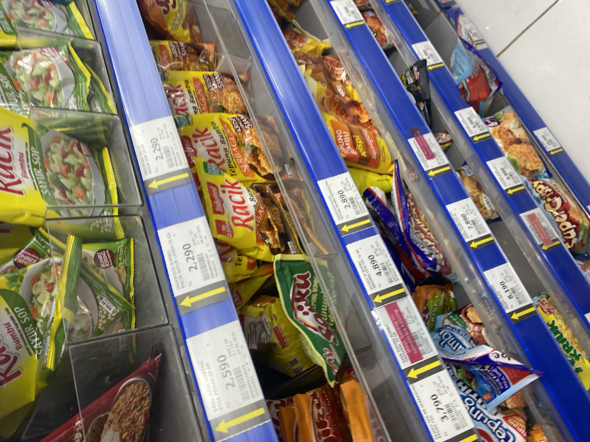 The image shows a retail environment, specifically a shelf in a supermarket or minimarket in Watampone, Indonesia. The shelves are stocked with various packaged food products, primarily seasonings and instant food mixes. Prominent brands visible include "Indofood Racik" and "Sasa Tepung Bumbu".
The products are displayed in clear plastic containers or directly on the blue-colored shelves. Each product appears to have a price tag attached to a yellow indicator arrow. Some of the visible product names include "Bumbu Sayur Sop", "Kobe Nasi Goreng Poll Pedas", "Sasa Tepung Bumbu Absorbs Less Oil", and "Tepung Bumbu Krispi Serbaguna".
The overall impression is a well-organized retail display focused on convenience food items. There are no people visible in the frame, and the focus is entirely on the products and their pricing. The lighting suggests an indoor setting typical of a commercial establishment. The image is taken at an angle looking down and across the shelves.