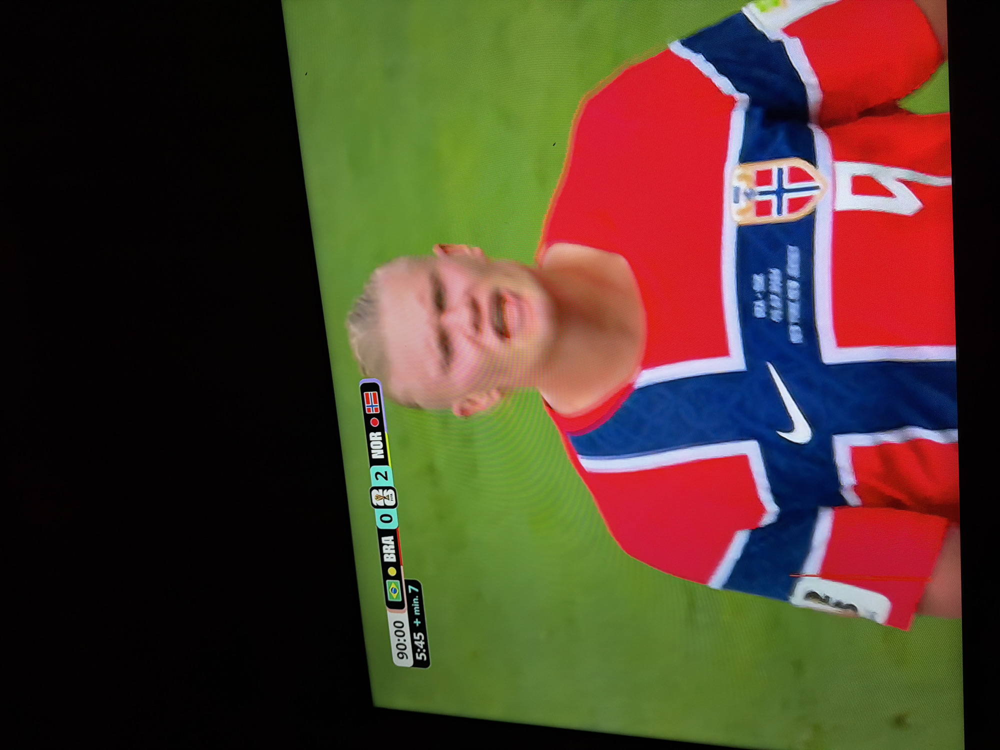 The image shows a close-up of a man wearing a red and blue soccer jersey with white trim. The jersey has a crest on the chest with a blue cross on a white background outlined in red, and what appears to be a red eagle above it. The man has blonde hair styled in a braid and is looking forward with his mouth open. A score graphic is visible in the upper left corner, indicating the time is 90:00 and the score is Brazil 0 - Norway 2, with an additional 5:45 + min. 7. The background is a blurred green field, consistent with a soccer pitch. The image is a photograph of a television screen, as evidenced by the slight glare and the pixelation. There is no indication of the city of Ognianovo, Bulgaria in the image.