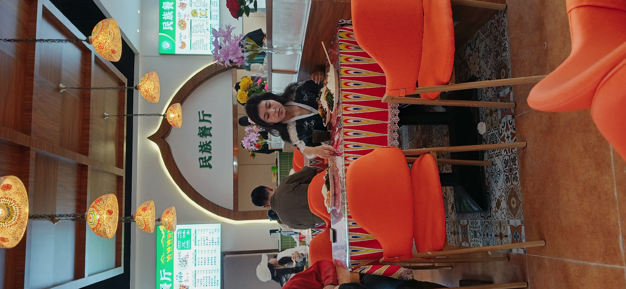 The image shows the interior of a restaurant located in Chongqing High-tech Zone, China. There are several patrons seated at a long table. A woman with dark hair is seated at the table and appears to be eating. Other people are seated nearby. The table has an ethnic-patterned runner. The restaurant has orange chairs. The décor includes ornamental hanging lamps and a sign with Chinese writing. Flowers are arranged on the table. A menu board is visible on the wall. The floor is tiled with a decorative pattern.