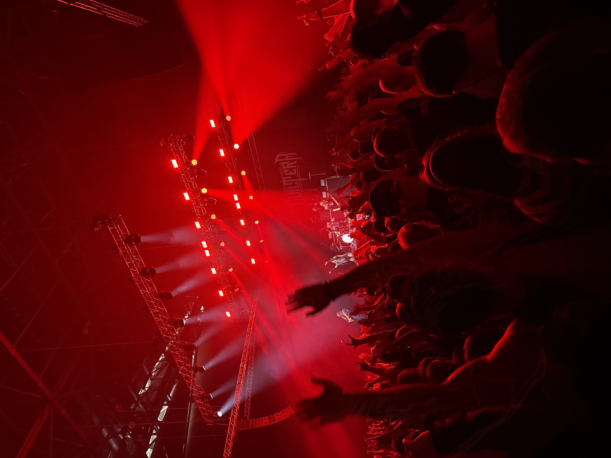 The image depicts a live concert setting, bathed predominantly in red light. On the left side, a metal stage structure with numerous light fixtures is visible. The right side of the image is filled with a crowd of people, many with their hands raised, likely reaching towards the stage. In the background, a logo of the band "PANTERA" can be seen. Given the information that this is in Sofia, Bulgaria, the concert is likely taking place at a large event venue such as the Arena Sofia. The actions portrayed are those of fans enjoying a live music performance.