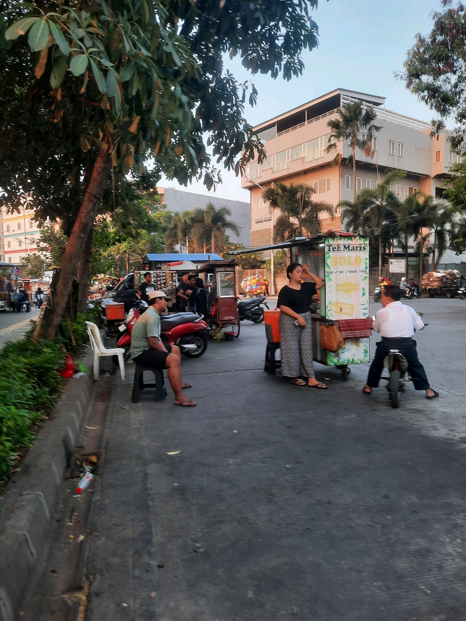 The image depicts a street scene in North Jakarta, Indonesia, likely during the late afternoon or early evening, given the lighting conditions. In the foreground, a man wearing a green t-shirt and dark shorts sits on a black stool next to a red motorcycle. He has a light-colored cap on and is looking to his right. To his left, a white plastic chair sits by a curb with a red plastic bag beside it.
Behind the man, a food cart is visible, displaying a sign that reads "Teh Maris SOLO" in large lettering. A woman in a black shirt and patterned pants stands next to the cart, appearing to interact with a man on a small motorcycle. The man on the motorcycle is wearing a white shirt and dark pants and is looking towards the cart.
Further down the street, several other motorcycles and a food cart are present. In the background, modern buildings with multiple stories and windows are visible, along with palm trees and other foliage. The street surface is asphalt, and there is a concrete curb with a drainage ditch. The overall impression is of a bustling urban environment with informal commerce and transportation.