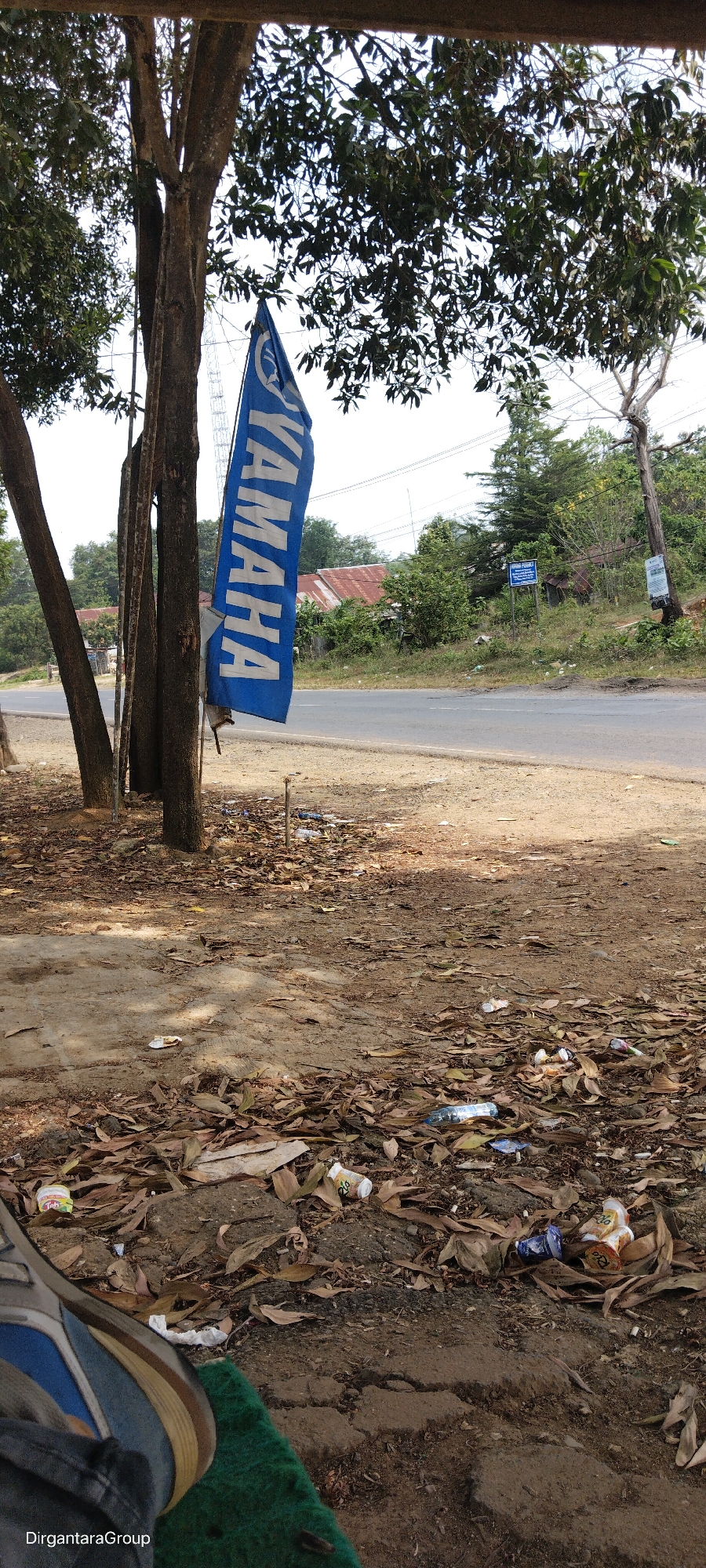 The image shows a Yamaha flag hanging from a tree next to a road. The flag is blue with white lettering that says "YAMAHA". The road is asphalt and appears to be in a rural area. There is some litter on the ground around the flag, including fallen leaves and plastic bottles. In the background, there are trees and some buildings with red roofs. There is a sign on a pole in the background with text on it, but it is too blurry to read. There is also a person's foot in the lower left corner of the image, wearing a sneaker and resting on a green mat. The overall scene suggests an outdoor location in a somewhat unkempt area.