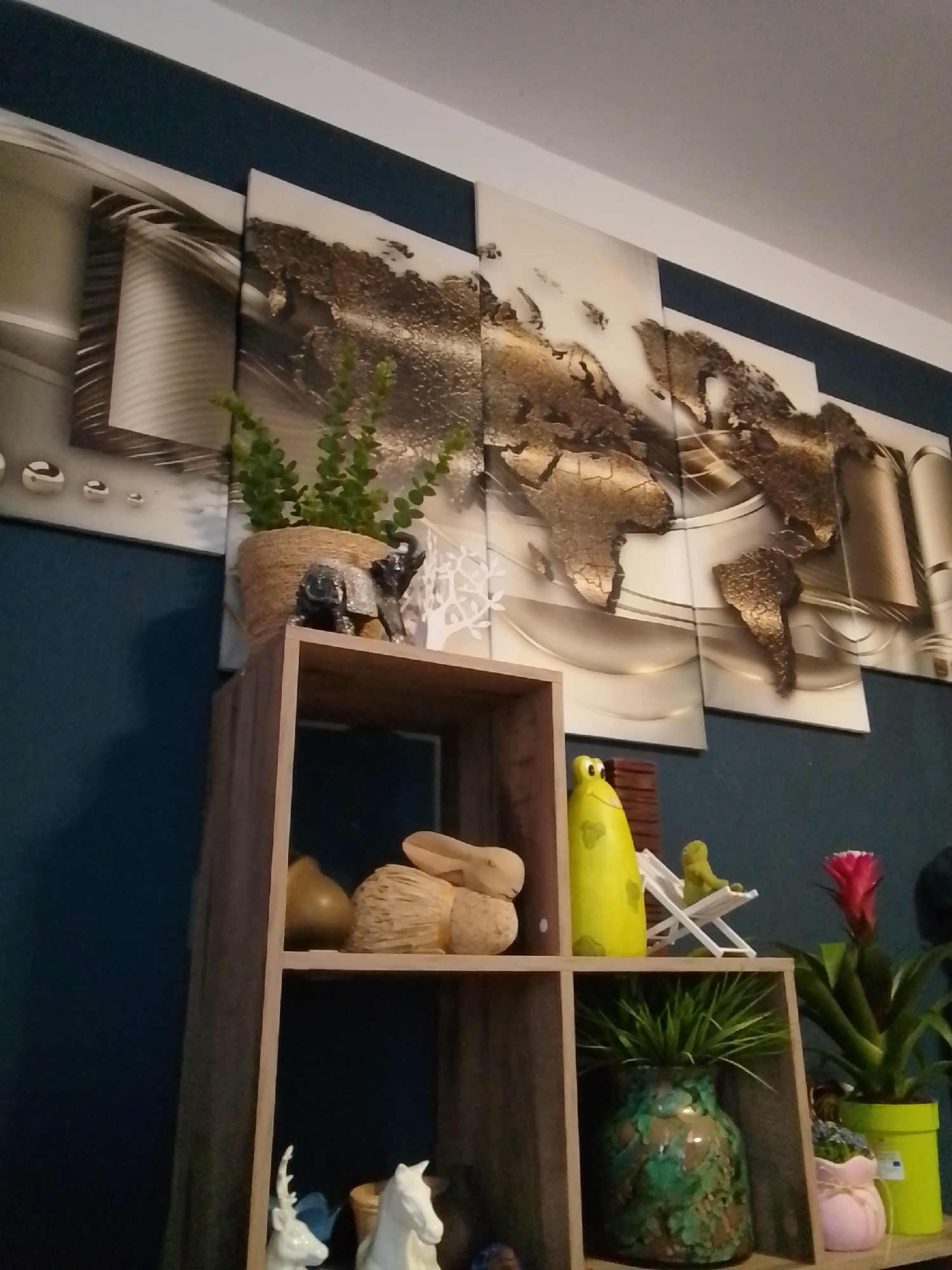 The image shows a shelf with decorative items, positioned against a dark blue wall. Above the shelf, a large, multi-panel wall art depicts a world map. The shelf itself is made of wood and has multiple compartments. On the top shelf, a potted plant sits next to a small, glittery elephant figurine and a white decorative tree. The middle shelf holds several decorative objects, including a yellow figurine of a frog, a small green frog figurine on a white lounge chair, and a pink flower in a green pot. The lower shelf contains a large green patterned vase with a plant, and to its right, a small pink pot with a light pink ribbon. On the bottom left of the shelf, there are white figurines of a deer and a horse. The scene appears to be an indoor home setting. The image does not provide any information or context that would allow for determining its location in the city of Wedel, Germany.
