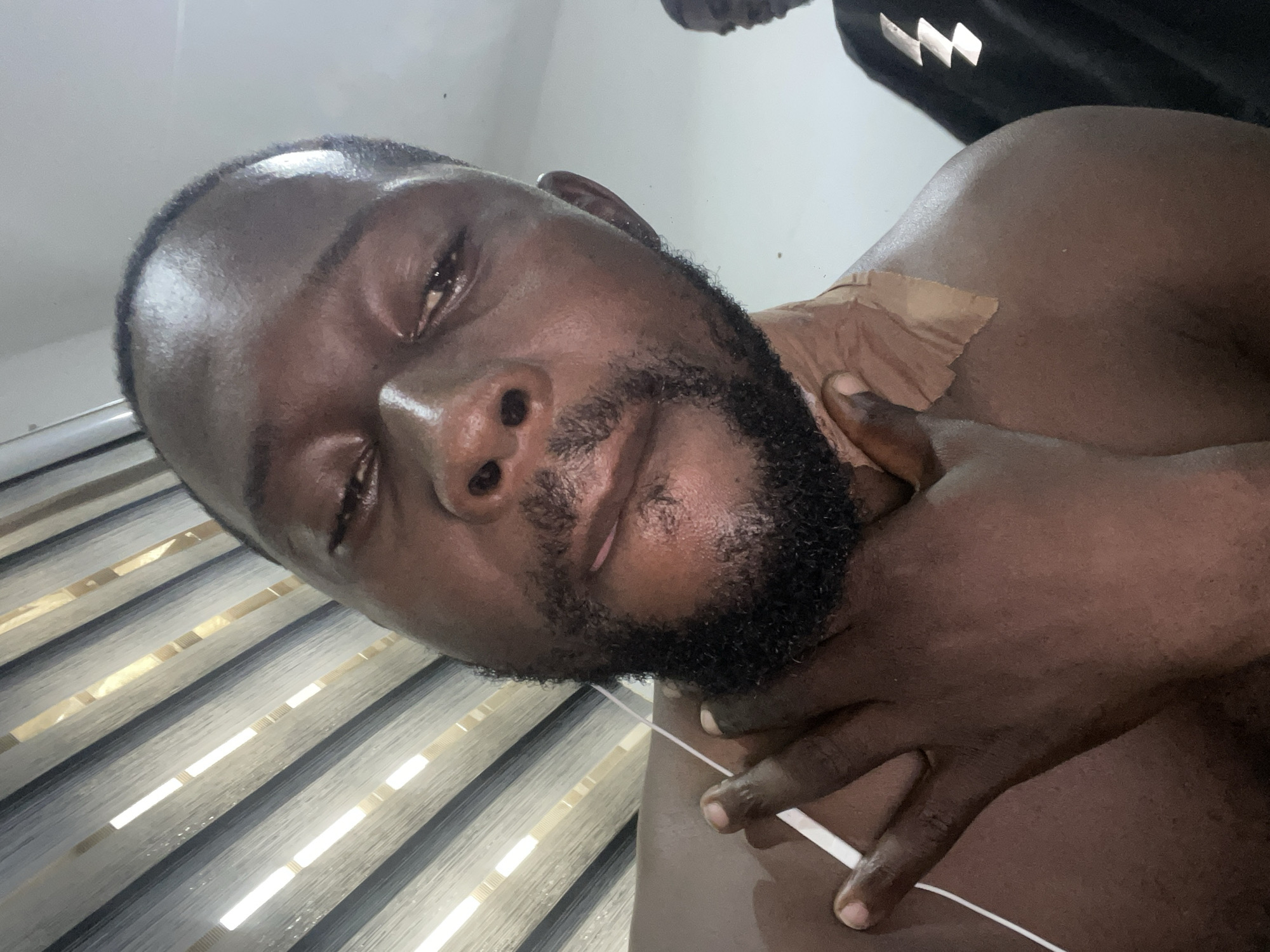 The image shows a close-up, low-angle, selfie-style shot of a man's face and upper body. He has dark skin, a beard, and appears to be wearing headphones with a white wire visible. His right hand is touching his neck, and there is a piece of light-brown medical tape on his shoulder. The background consists of horizontal blinds with light shining through them. To the right, another person in a black garment with a white lightning bolt design is partially visible. The scene appears to be indoors, possibly in a residential setting within the city of Abuja, Nigeria.