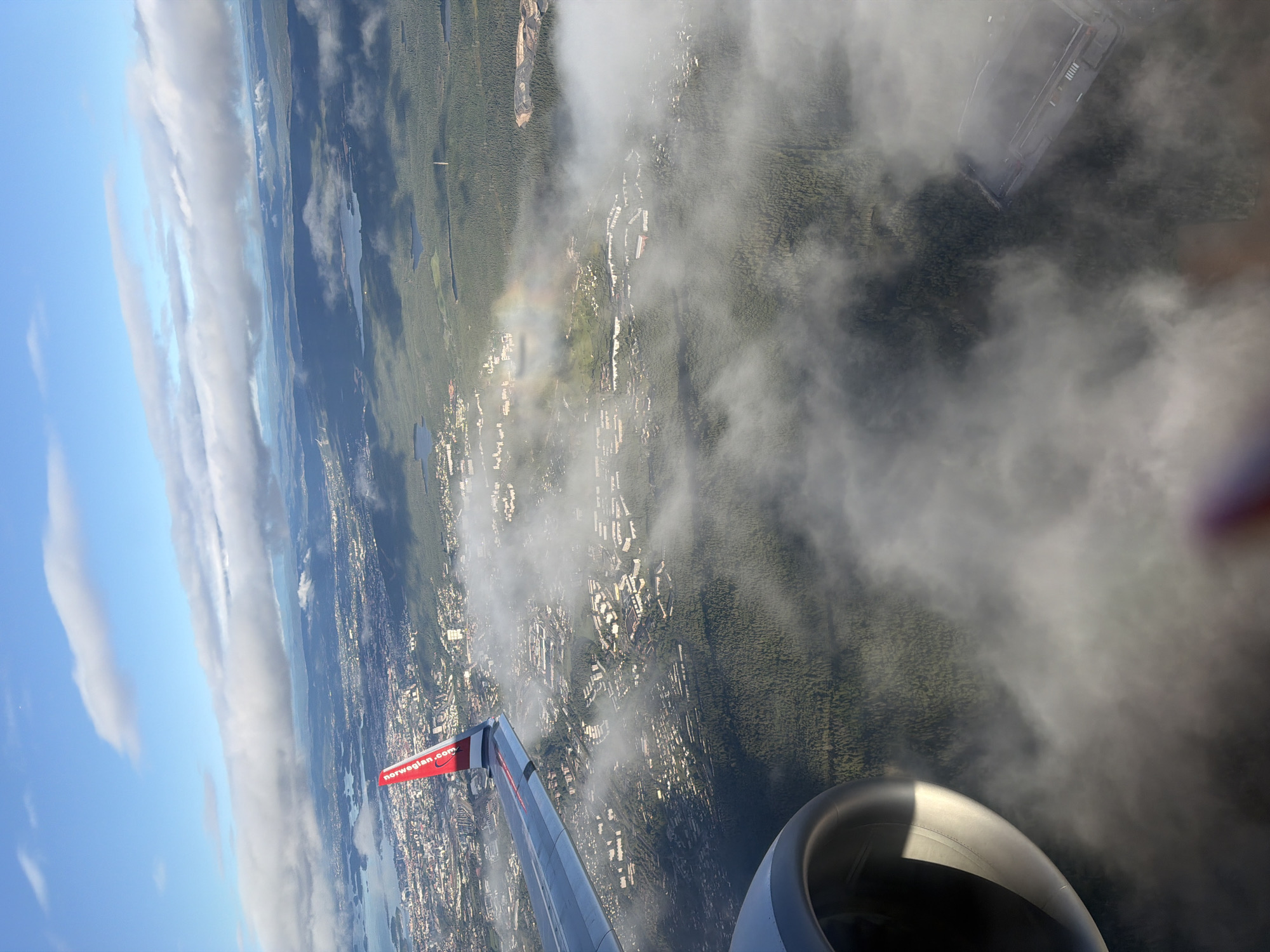 The image is an aerial view from an airplane window, looking down on Gardermoen, Norway. A portion of the airplane's wing, with the red and white "norwegian.com" logo, is visible in the upper left quadrant. Below the wing, a sprawling urban area with dense housing and a significant amount of green forest cover can be seen. Patches of fog or clouds are scattered across the landscape, partially obscuring the view of the city and the terrain. A faint rainbow is visible in the distance, amidst the clouds and over a built-up area. In the lower left corner of the image, the engine nacelle of the aircraft is prominent. Further into the landscape, there are glimpses of water bodies, and a large, rectangular structure that appears to be a facility or building complex is visible towards the bottom right, partially shrouded by fog. The overall scene depicts a partially cloudy day over a Scandinavian city and its surrounding natural environment.