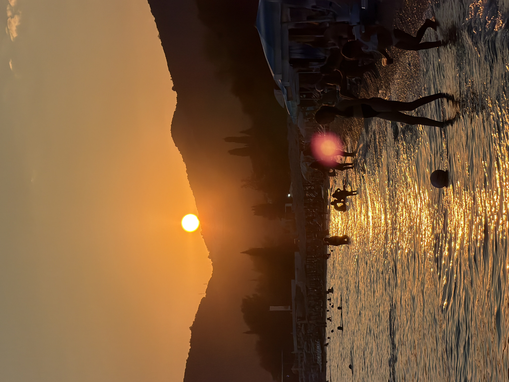 The image depicts a sunset scene at a beach in Boreti, Montenegro. Silhouettes of people are visible in the shallow water and on the shore. Two children in the foreground appear to be playing with a ball in the water. In the background, there are hills, trees, and beach umbrellas, all cast in the warm, golden light of the setting sun. The sun itself is a bright, luminous orb positioned just above the horizon, casting a shimmering reflection on the water's surface. The overall atmosphere is one of a serene and picturesque evening at the seaside.