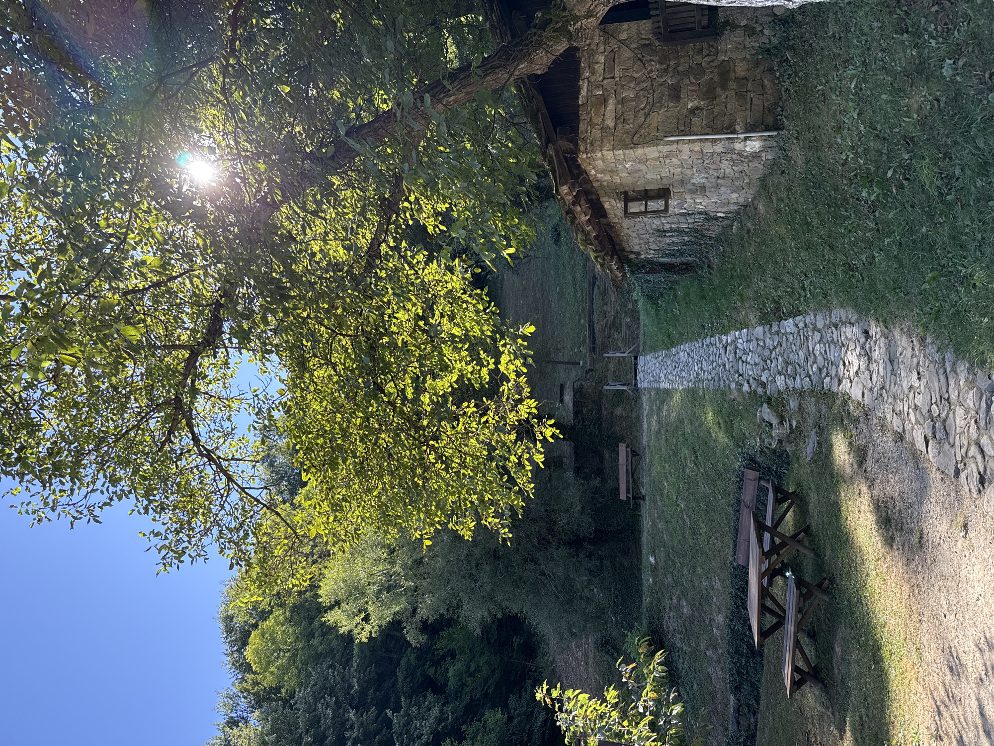 The image depicts an outdoor scene with a stone-built structure and a path leading towards it. The stone building, located on the right side of the image, has a rustic appearance with a moss-covered roof and a small, wooden-framed window. A cobbled path runs from the foreground towards the building, winding through a grassy area.
In the foreground, to the left, there is a wooden picnic table with two benches. Sunlight filters through the lush green leaves of a large tree that arches over the scene, casting dappled shadows on the ground and path. Behind the picnic table and further up the slope, another wooden bench is visible.
The background is dominated by dense green foliage, suggesting a wooded or hilly environment. The sky is a clear, bright blue. The overall impression is one of a tranquil, natural setting, possibly a rural retreat or a park area.
This scene is located at the Etar Architectural-Ethnographic Complex, a museum in Gabrovo, Bulgaria, that showcases traditional Bulgarian crafts and rural architecture. The stone building is representative of the historical houses found in the complex, and the picnic areas and paths are designed to enhance the visitor experience.