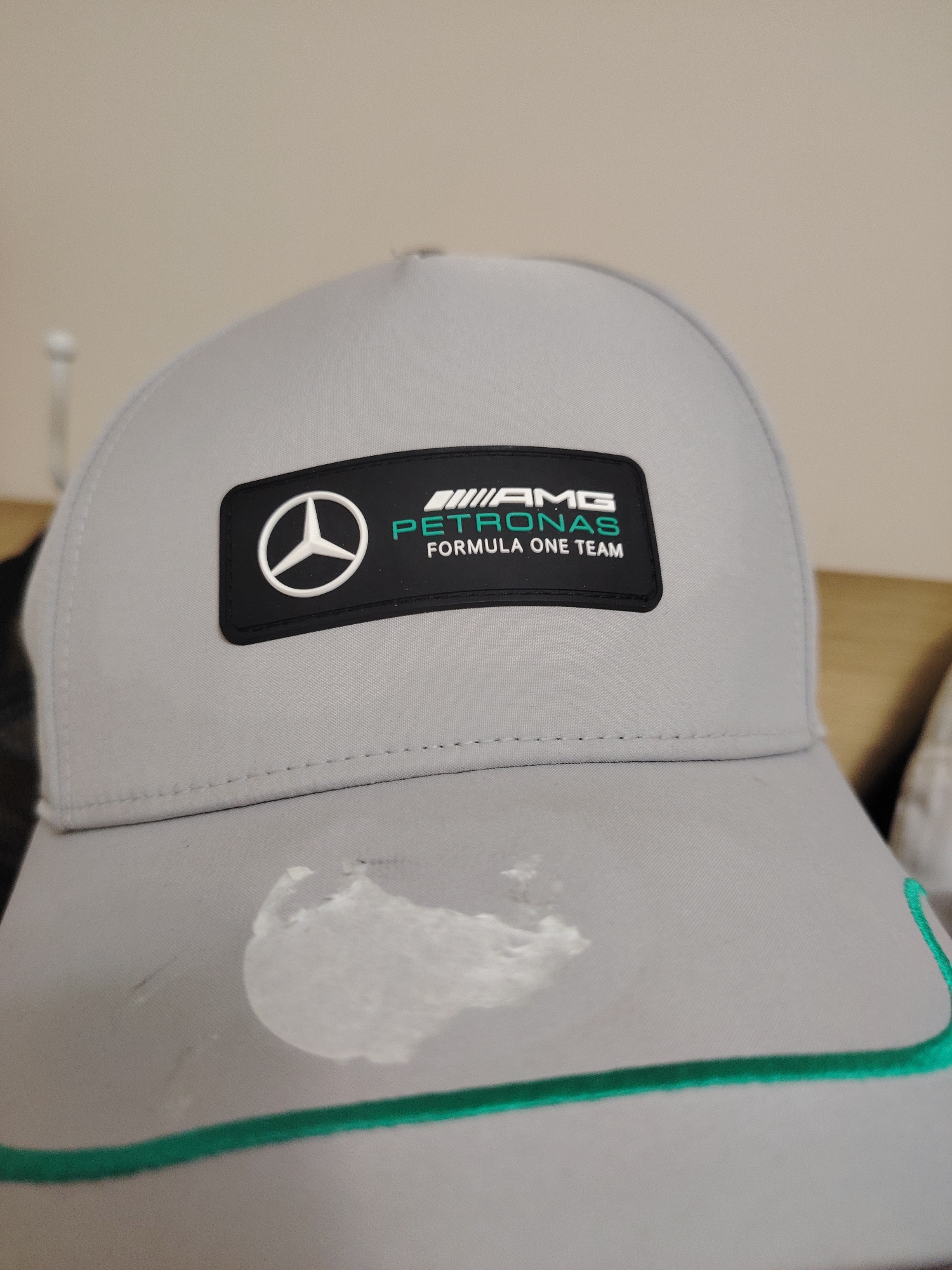 The image displays a close-up, slightly angled view of the front of a light grey baseball cap. Centered on the cap's front panel is a black rectangular patch with subtly rounded corners. On the left side of this patch, a white, raised Mercedes-Benz three-pointed star logo is featured. To the right, white text reads "///AMG" above teal-green text "PETRONAS", which is further above white text "FORMULA ONE TEAM". Horizontal stitching lines are visible on the cap's fabric above the brim. The front edge of the cap's brim is adorned with a narrow, embroidered teal-green line. A significant, irregular white mark, consistent with dried residue or a substance, is visible on the cap's main panel below the black emblem, accompanied by smaller, fainter smudges to its left. The background consists primarily of a plain, light beige wall, with a hint of a brown wooden surface visible to the right.