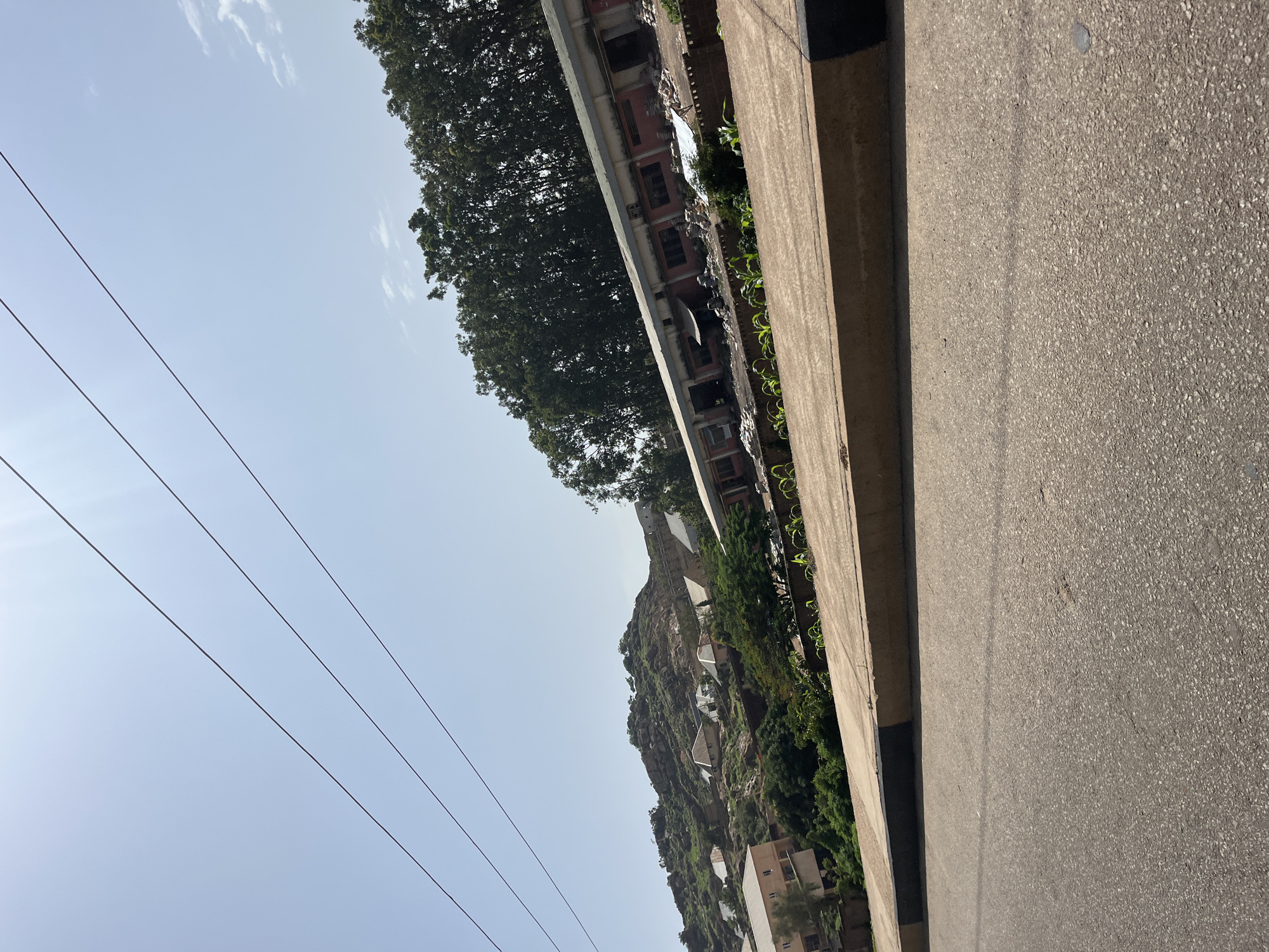 The image depicts a street-level view in Gura Top, Nigeria, on a clear, sunny day. In the foreground, a paved road stretches into the distance, with a concrete curb bordering its right side, marked with black and yellow stripes. Beyond the curb, a line of young green plants is visible.
In the mid-ground, a long, low building with a reddish-pink facade and a metallic roof runs horizontally across the frame. This building appears to be industrial or commercial, with several doorways and windows. Debris and piles of materials are scattered in front of it. Behind the building, a dense line of tall, leafy green trees forms a natural barrier.
The background features a sloping hillside covered in rocks and sparse vegetation. Several houses with white and light-colored roofs are scattered across the hillside, indicating a residential area built into the terrain. Three thin, dark power lines stretch diagonally across the clear blue sky, originating from the left side of the frame and extending towards the upper right. The overall impression is of a somewhat arid, hilly landscape with developing infrastructure.