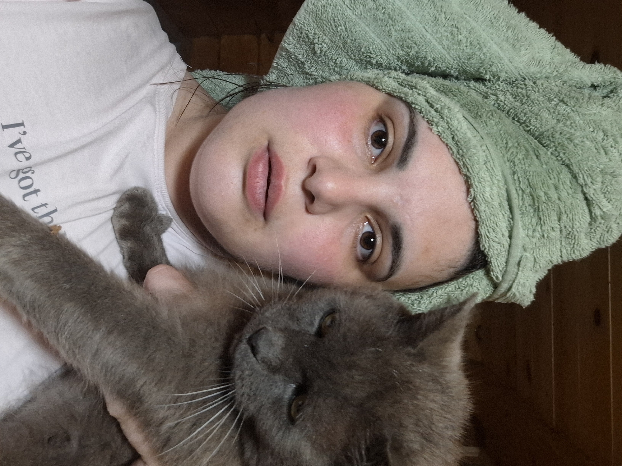 The image shows a person and a gray cat, possibly inside a home or building in the city of Амзя, Russia. The person is wearing a white t-shirt and has a green towel wrapped around their head. The cat is being held by the person. The cat's fur is gray, and it has white whiskers.