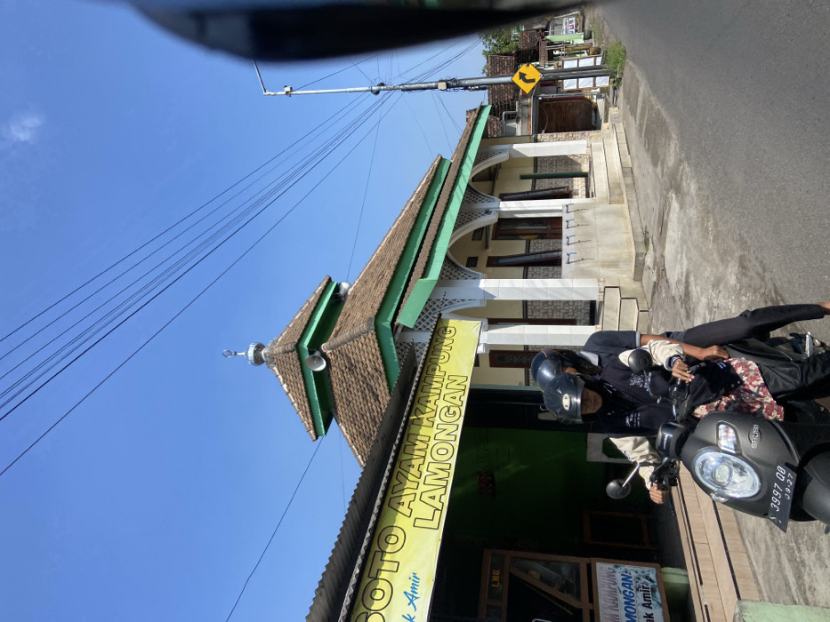 The image shows a street scene in Mojosari, Indonesia, located at latitude -7.503 and longitude 112.543. A mosque with a green roof and white walls is located on the right side of the street. There are power lines running above the street. The sign on the left side of the street reads "Soto Ayam Kampung Lamongan," which likely indicates a restaurant. Two people, one male and one female, are riding a black scooter in the street. The female is wearing a black hijab and a black jacket, while the male is wearing a black helmet and a blue and black jacket. The scooter is facing the viewer, and the male is holding the handlebars. The female is sitting behind the male, and her hands are on the scooter's handlebars. The scooter has a license plate that reads "3997 DD." The scene is bright and sunny, and the sky is blue. There are multiple buildings on the left and right of the street. There is also a sign with a yellow background and a curved arrow on the right side of the street, indicating a turn. The street is paved and has a white curb. The image is taken from a slightly elevated position, possibly from a car. The scene is quiet and peaceful.