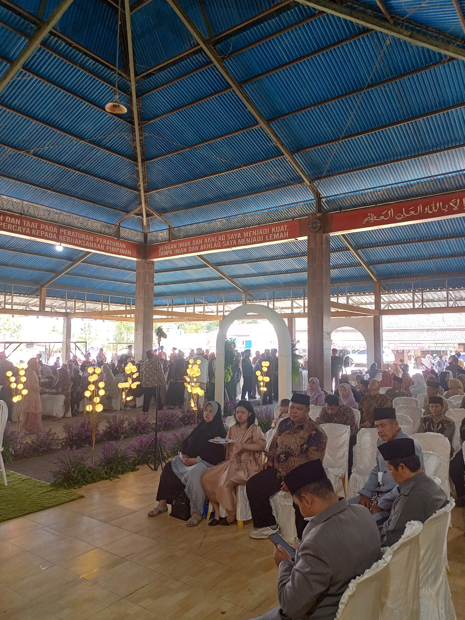 The image depicts an indoor gathering in Wanadadi, Indonesia, likely at a community hall or event space. The ceiling is constructed of corrugated blue metal supported by exposed metal beams and a central concrete pillar. Red banners with white text are visible along the upper walls. One banner displays Arabic script, and another contains Indonesian text referencing adherence to rules and leadership.
A large group of people are seated on white chairs arranged in rows, facing an unseen focal point. Many are dressed in traditional Indonesian attire, including peci hats and batik patterns. Some individuals are looking towards the front, while others are engaged in conversation or looking at their phones. In the foreground, several people are seated, and a man in a grey suit is looking at his mobile phone. To the left, decorative yellow globe lights on tall stalks are placed amongst purple flowers, creating a festive ambiance. An arched opening with a white frame is visible in the background, leading to another part of the venue where more people are gathered. The floor is tiled with light-colored wooden planks. The overall atmosphere appears to be one of a formal occasion, possibly a religious or community event.