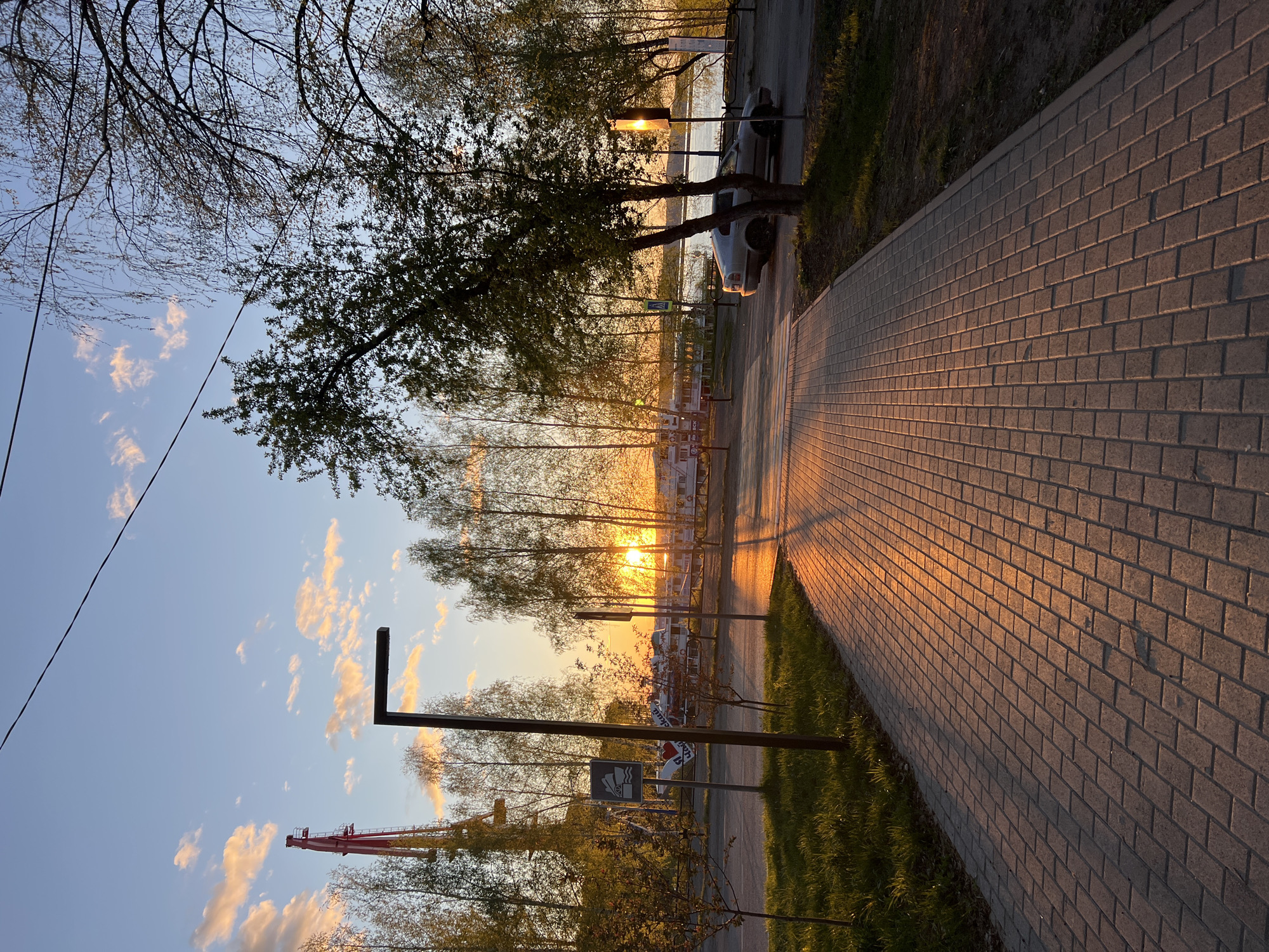 The image captures a sunset scene in Chaikovsky, Russia. A brick pathway dominates the foreground, angling from the lower left to the upper right. The setting sun casts long shadows across the path, creating an orange glow. To the right of the path, a narrow strip of grass borders the edge. On the left side of the path, a row of trees lines the road. Beyond the trees, the setting sun is visible, illuminating the sky and the river with a golden hue. A car is parked alongside the road among the trees. A red construction crane is partially visible in the lower left of the image. A few clouds are scattered across the blue sky. A street light is present.