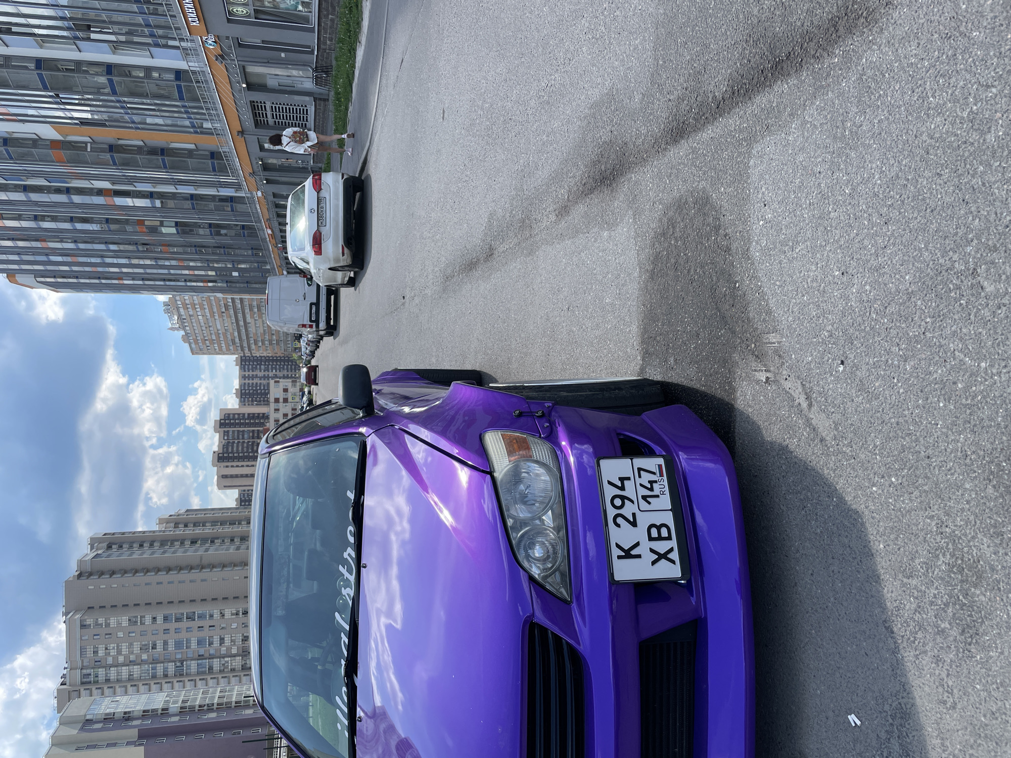 The image captures a sunlit urban street scene in the city of Unknown, Russia. A highly customized, bright purple sedan, identified by Russian license plate K 294 XB 147 RUS, is prominently parked on the left side, partially obscuring the front of the vehicle. It features aftermarket wide fender flares, a pronounced front splitter, and a windshield banner reading "illegal street."
The asphalt road, showing dark linear tire marks or stains, extends into the distance. On the right side of the street, a white Volkswagen sedan, bearing Russian license plate M 380 KB 196 RUS, is parked further along, facing away from the camera. Behind it, a white panel van is also parked. A single pedestrian is visible on the sidewalk to the right, walking away from the viewer, dressed in a white shirt, shorts, and carrying a small backpack.
The architecture in the background consists of numerous modern, multi-story buildings. On the left, several high-rise residential structures with light-colored and purple accents are visible. On the right, a building with a contemporary facade of glass, grey, and orange panels houses ground-floor businesses, one partially identified by the Cyrillic word "КЛИНИК" (Klinik - Clinic). The sky overhead is blue with scattered white cumulus clouds, indicating clear daytime conditions and casting visible shadows on the pavement.