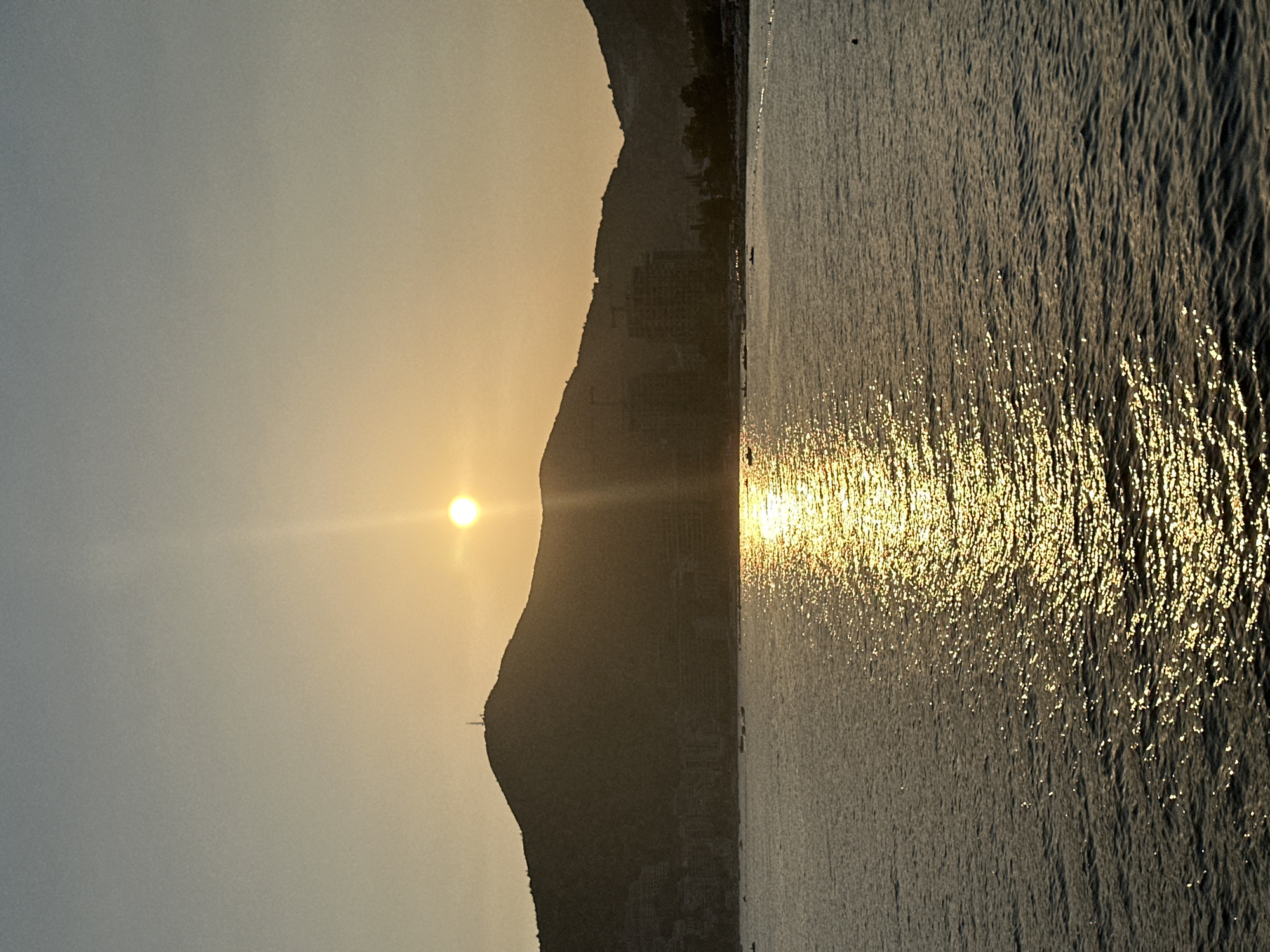The image depicts a serene sunset over the Adriatic Sea in Budva, Montenegro. The sun, a bright orb, is positioned in the upper center of the frame, casting a golden reflection across the water. In the background, silhouetted against the warm hues of the sky, are the lush, rolling hills that surround the city. The slopes of the hills are dotted with buildings, suggesting the urban landscape of Budva is nestled against the natural topography. A prominent tower, likely a communication mast, stands atop the highest peak. The sea is calm, with gentle ripples creating a shimmering effect as the sunlight plays on its surface. Several small boats are visible on the water, adding a sense of human activity to the tranquil scene. The overall atmosphere is one of peaceful transition from day to night, with the natural beauty of Montenegro's coastline taking center stage.