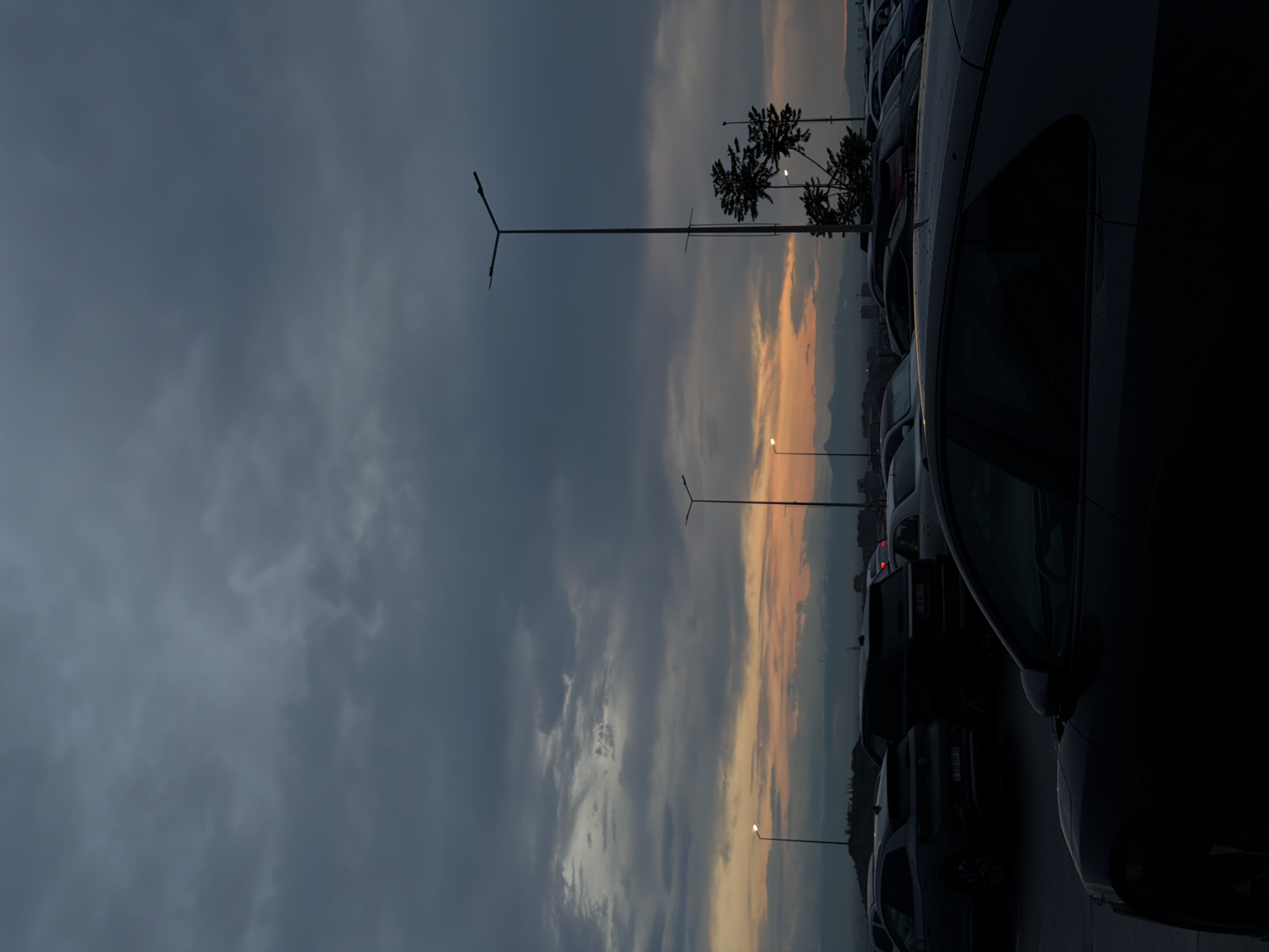The image depicts a parking lot in Yerevan, Armenia, under a dramatic, overcast sky. The sky transitions from a dark, cloudy gray to a vivid orange and yellow near the horizon, suggesting either sunrise or sunset. Rows of parked cars fill the right side of the frame, with the nearest car appearing quite dark and dominating the foreground. Streetlights are visible amidst the parked vehicles, their lamps illuminated against the twilight. A few trees are silhouetted against the lighter sky. In the distance, a faint outline of the city skyline can be observed.