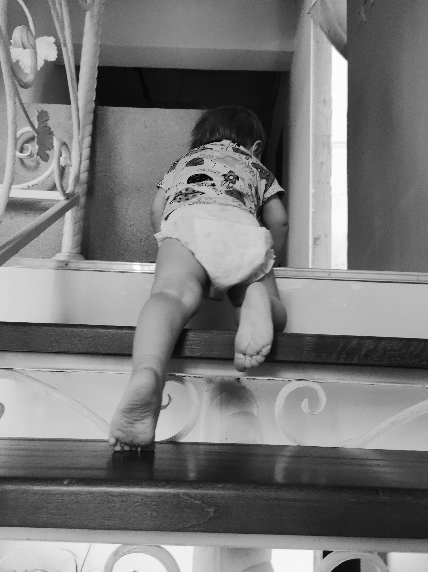 A black-and-white photograph captures a toddler climbing a staircase. The child, seen from behind, is wearing a patterned shirt and a diaper. The child's bare feet are visible as they step on the wooden steps. The staircase has a decorative metal railing with ornate designs. A doorway is visible at the top of the stairs. The specific location is unidentified.