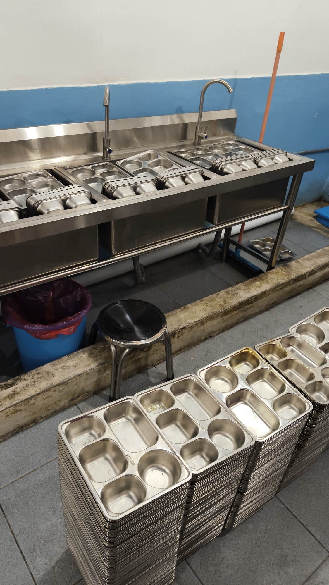 In Sambipondok, Indonesia, an industrial kitchen is depicted, featuring a stainless steel counter with multiple food service trays. Two faucets are visible on the counter, suggesting a dishwashing or food preparation area. Below the counter, a blue trash bin with a purple liner is situated, alongside a small, round black stool. In the foreground, stacks of metal food trays are neatly arranged, indicating preparation for service or storage. The environment appears clean and organized, consistent with a food service establishment.