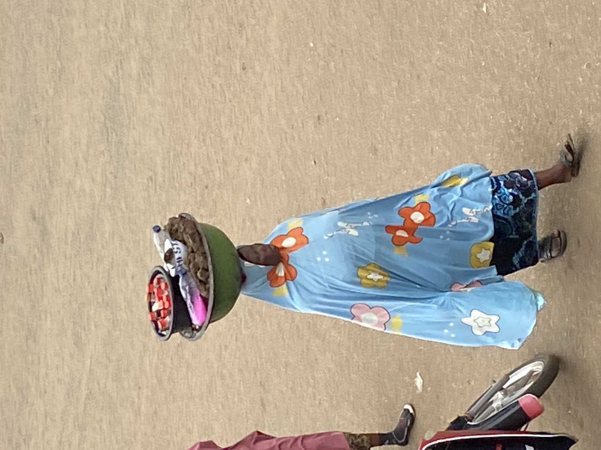 A street scene in Bukuru, Nigeria. A woman in a blue floral dress balances a bowl of goods on her head. A motorcycle is nearby. Another person’s legs are visible in the lower left. The setting appears to be a street.