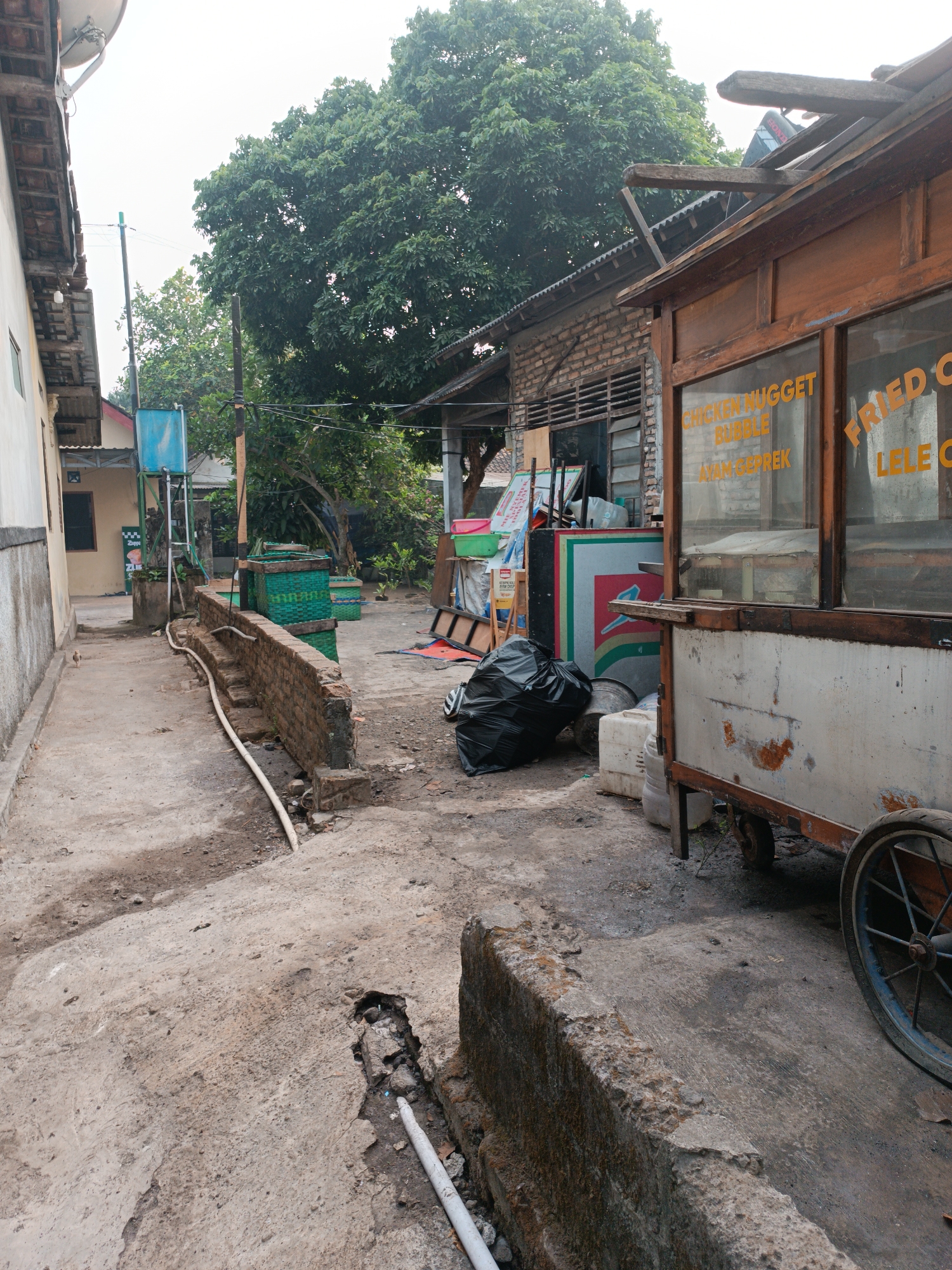 An outdoor scene in Jetis, Indonesia, depicts a narrow concrete pathway flanked by buildings. To the left, a pale yellow wall of a building runs along the path, with a satellite dish mounted on its roof. On the right, a wooden food cart with a weathered exterior and a window displays text reading "CHICKEN NUGGET", "BUBBLE", and "AYAM GEPREK" on one side, and "FRIED C" and "LELE C" on the other. The cart is positioned on a rough concrete surface with visible dirt and debris. Behind the cart, a brick building with louvered windows and a corrugated metal roof is visible. A large green tree with dense foliage dominates the background. In the foreground, a broken concrete barrier reveals exposed rocks and a white pipe running along the ground. A black garbage bag rests near the food cart, and several green woven baskets are stacked in the middle ground. A small bird stands on the concrete pathway. The sky is overcast.