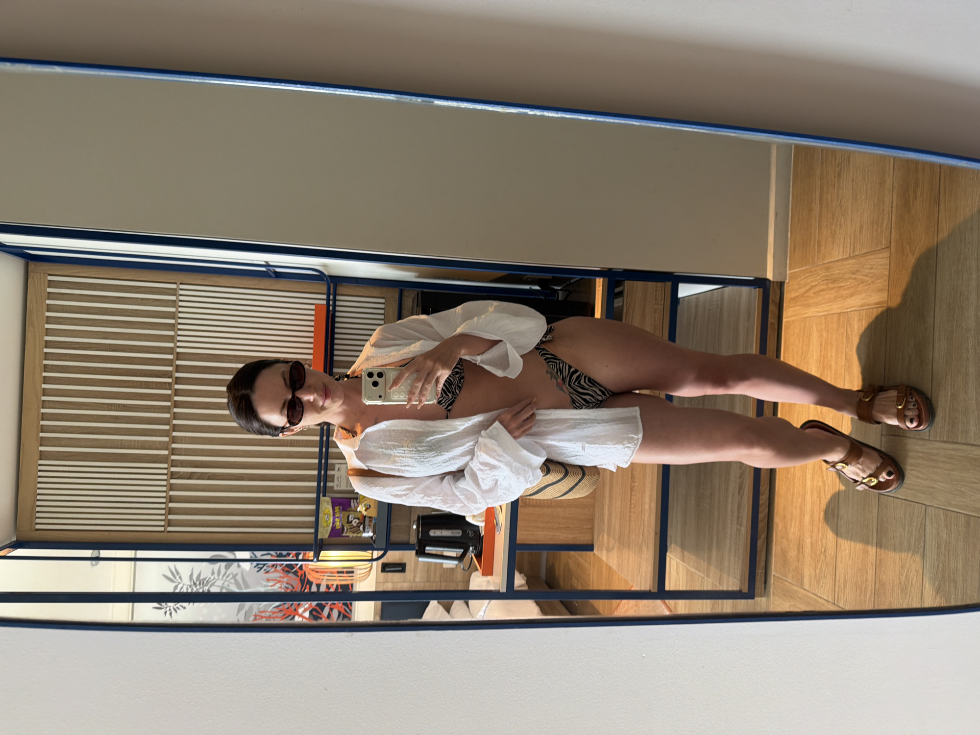 A woman is taking a mirror selfie in what appears to be a hotel room. She is wearing a white open button-down shirt over a black and white zebra-print bikini. She has on dark oval sunglasses and brown sandals. A straw tote bag is slung over her left shoulder. The room has light wood flooring and a wooden shelving unit with blue accents. A black electric kettle sits on the shelf. The overall impression is that of a resort or hotel setting. The provided information about the location in Dong Ninh Hoa Ward, Vietnam is not visually verifiable from the image itself.