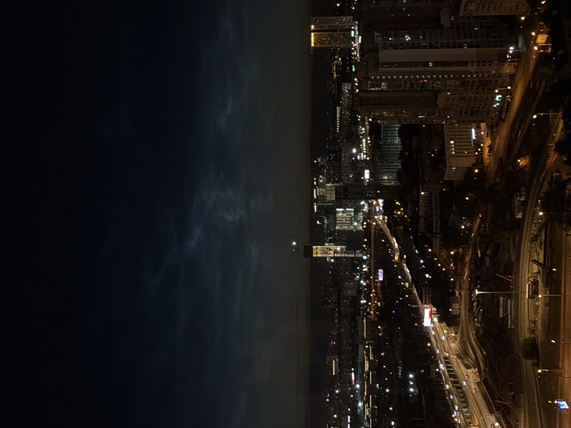 The image depicts a nighttime aerial view of the city of Khimki, Russia, showing a sprawling urban landscape illuminated by artificial lights. The sky is dark with subtle cloud formations and a faint glow near the horizon. In the mid-ground, a prominent tall skyscraper, lit with a yellow glow, stands out amongst other illuminated buildings of varying heights. A multi-lane highway with visible traffic, indicated by streaks of headlights and taillights, cuts through the scene, demonstrating ongoing urban activity. Construction sites with cranes and equipment are also visible, suggesting ongoing development in certain areas of the city. Streetlights and building lights create a dense network of illumination across the urban expanse, highlighting residential and commercial structures. The overall scene conveys a sense of a vibrant, active city at night.