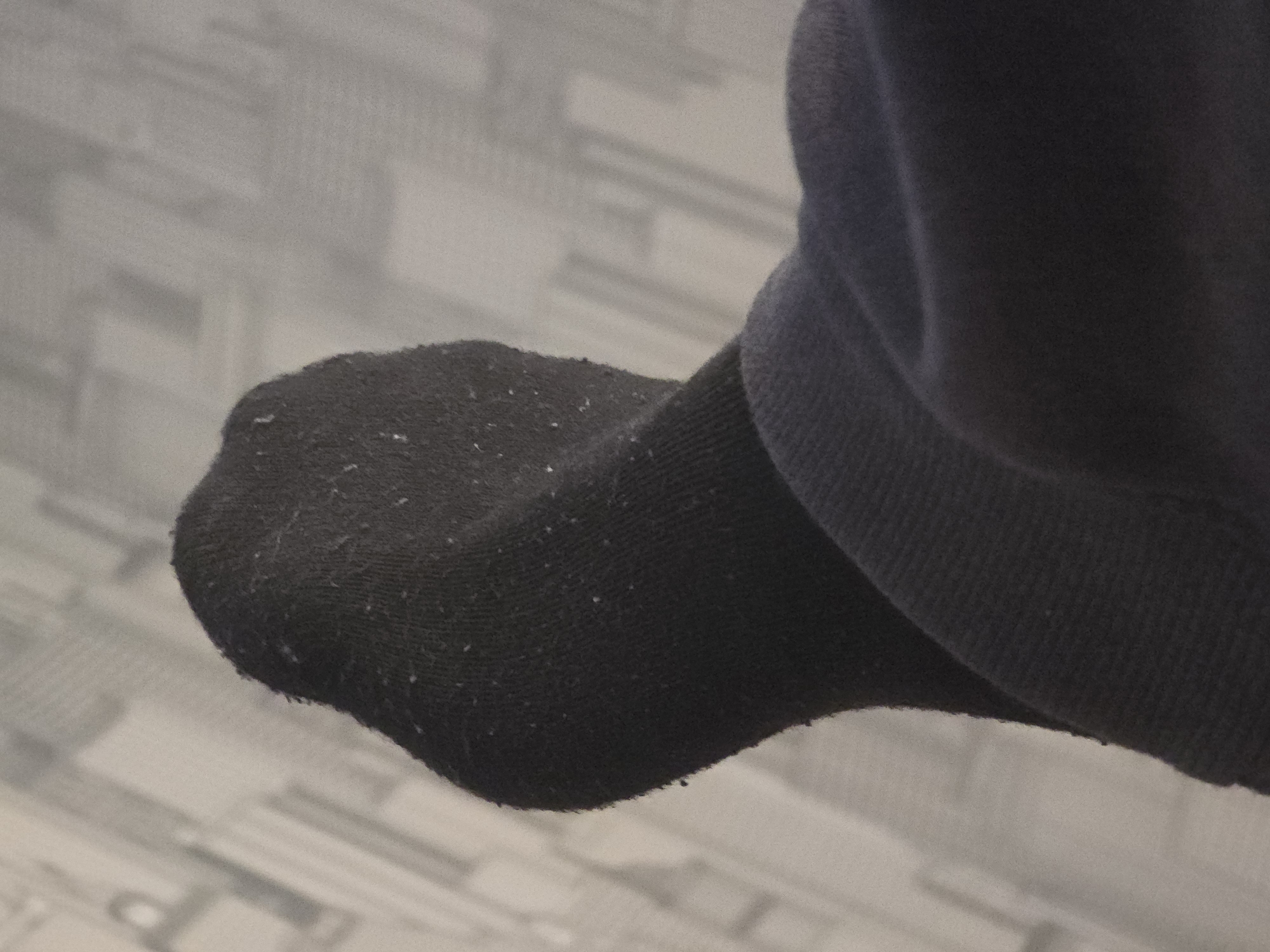 This media file features a close-up shot of a black sock, likely on a person's foot, resting against a textured, light-colored surface that appears to be a wall or fabric with a geometric pattern. The sock is dark and shows signs of wear, with small white specks, possibly lint or debris, visible on its surface, particularly around the toe area. The fabric of the sock has a knitted texture. To the right of the sock, a portion of dark fabric, possibly clothing like a sleeve or pant leg, is visible. The lighting in the image is soft, suggesting an indoor setting, and there are no clear indicators of time of day or weather conditions. The image is focused on the sock and its immediate surroundings, with the background being out of focus. No text is visible in the media file.